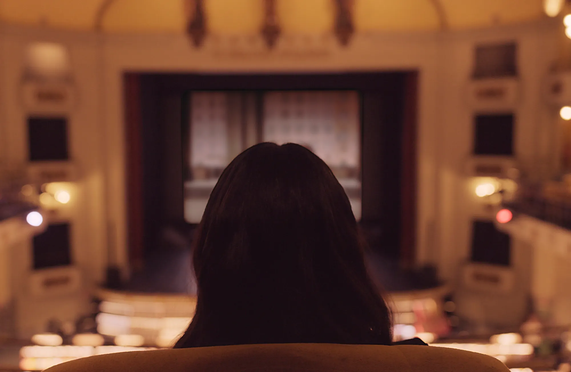 The back of a person with dark hair sitting in a cinema, facing a blurred movie screen. The warm-lit, classic architecture of the theater surrounds them in soft focus.