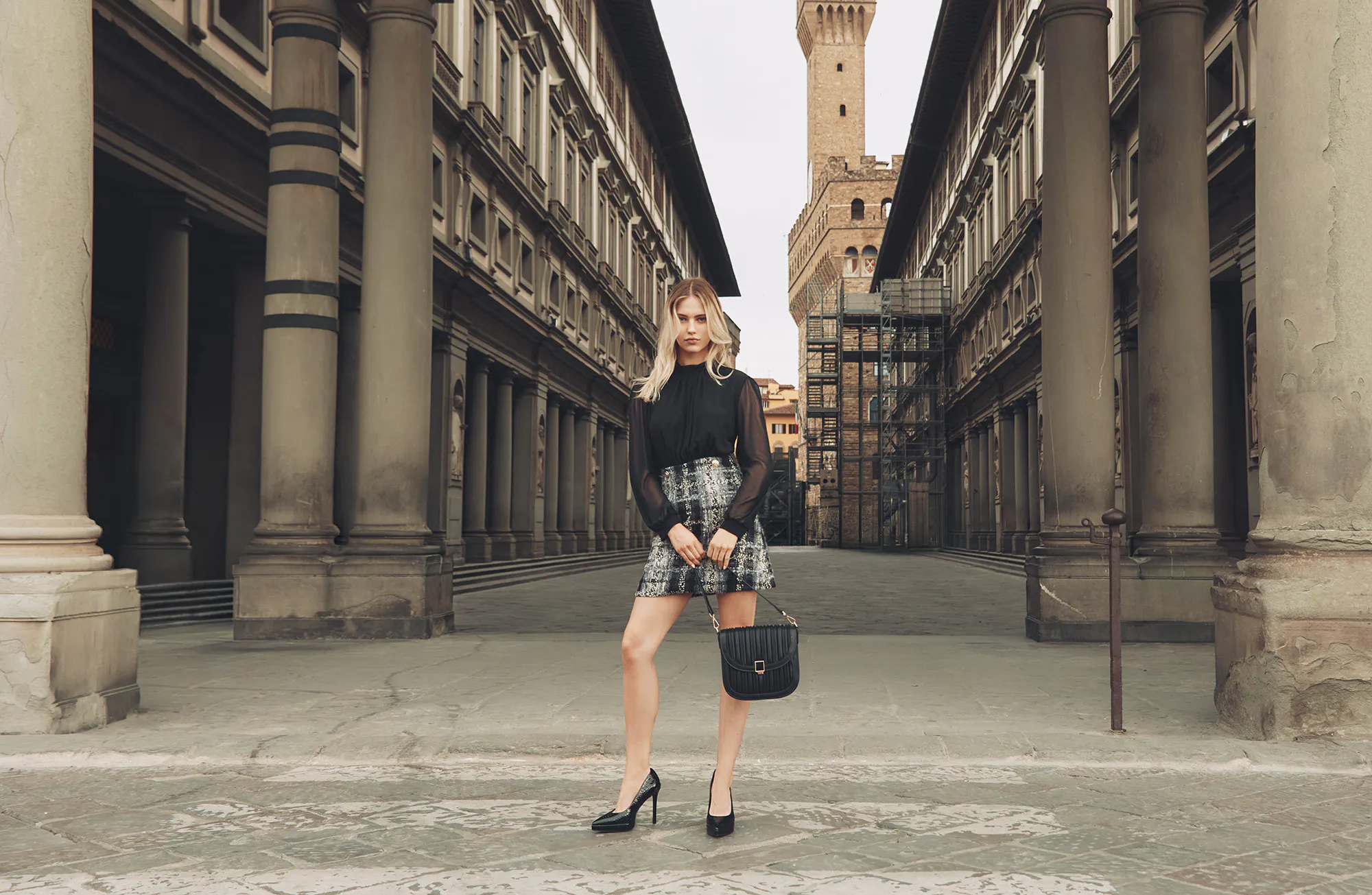 Young woman in a black top and patterned skirt poses with a handbag in a historic city square framed by tall columns and buildings.