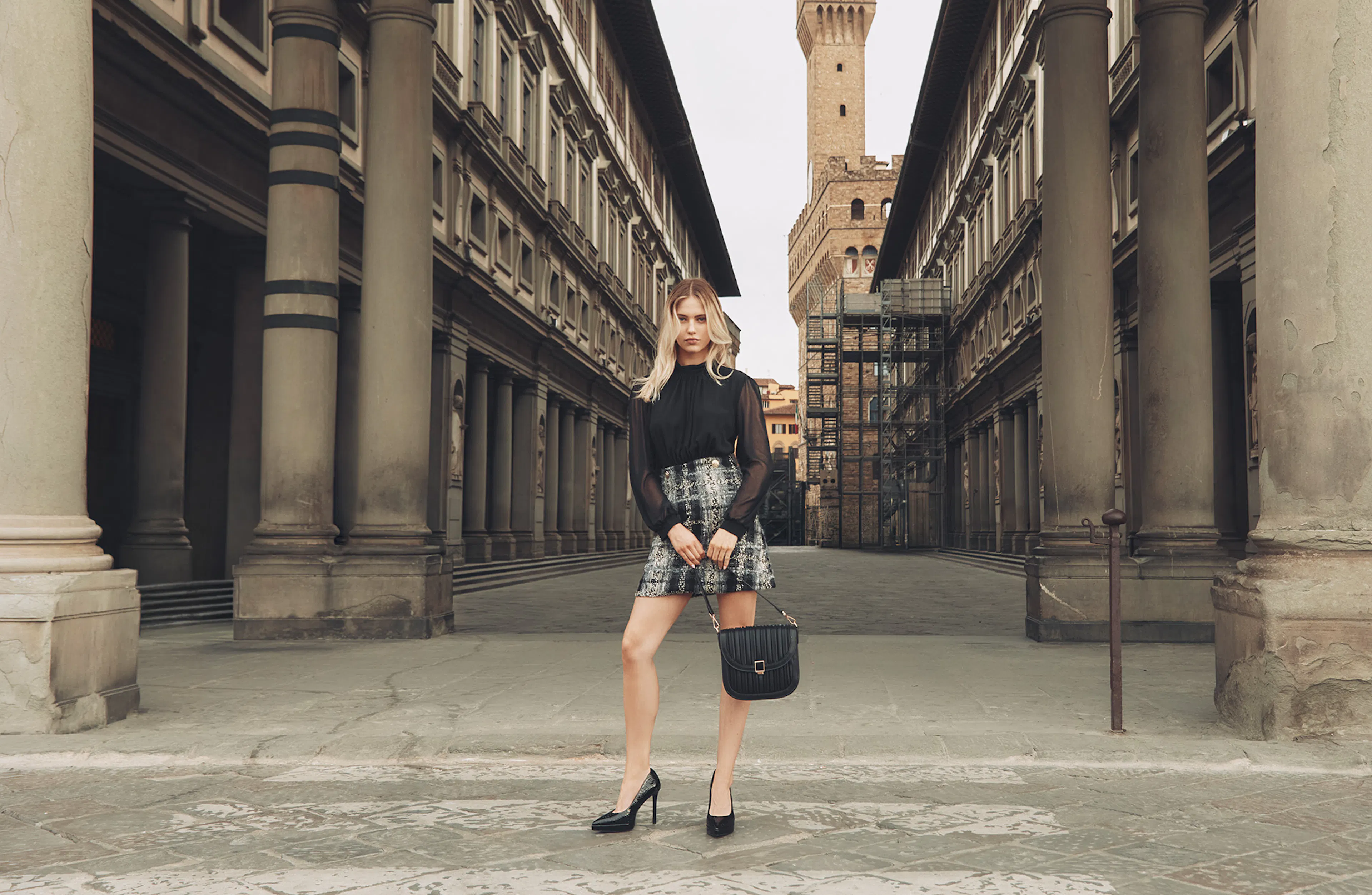 Young woman in a black top and patterned skirt poses with a handbag in a historic city square framed by tall columns and buildings.