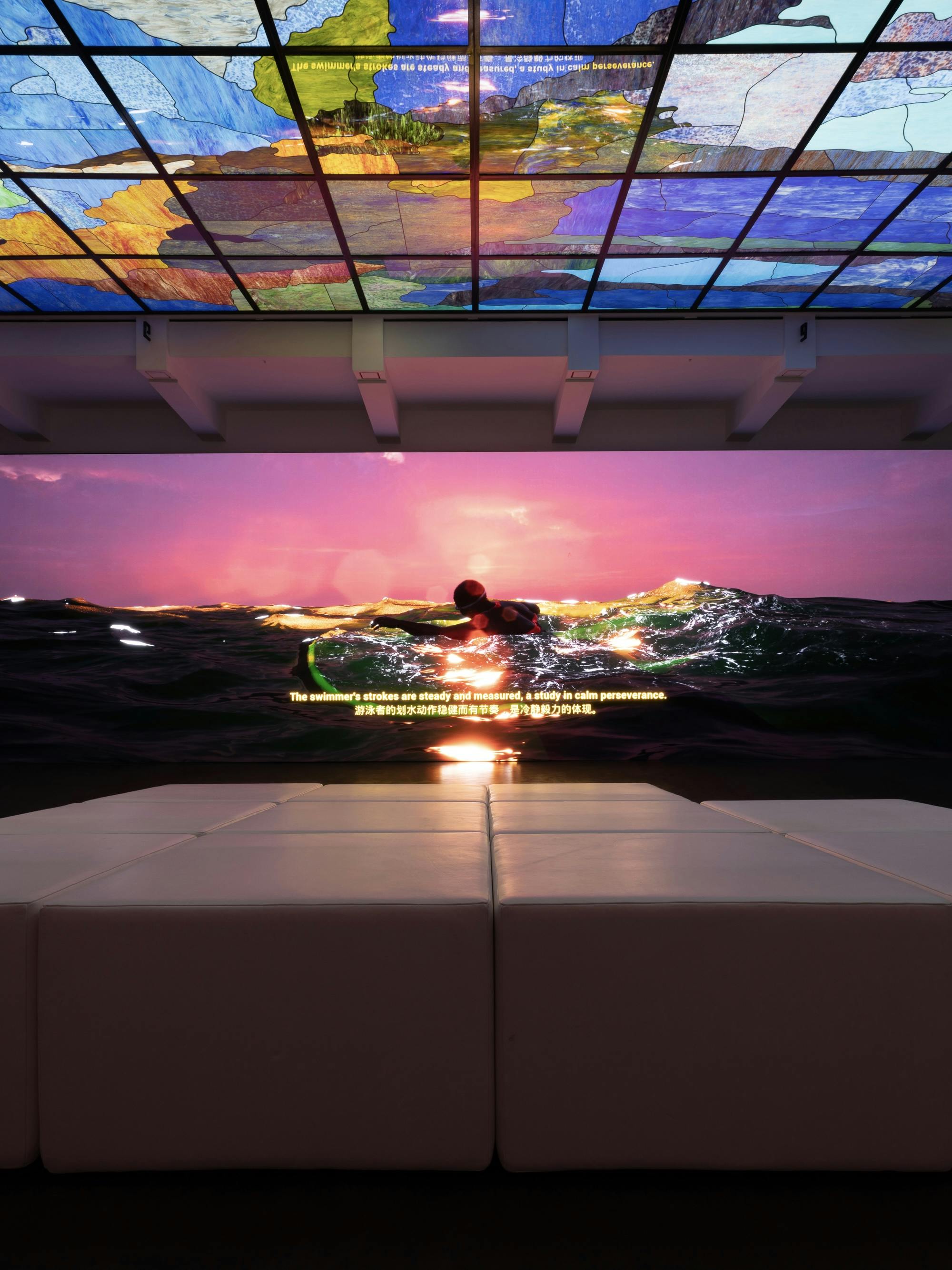 Center, a simulated animation of a person swimming through the pacific at sunset, in front is a slopped sofa shot from behind and above is a stained glass ceiling which is many different colors.