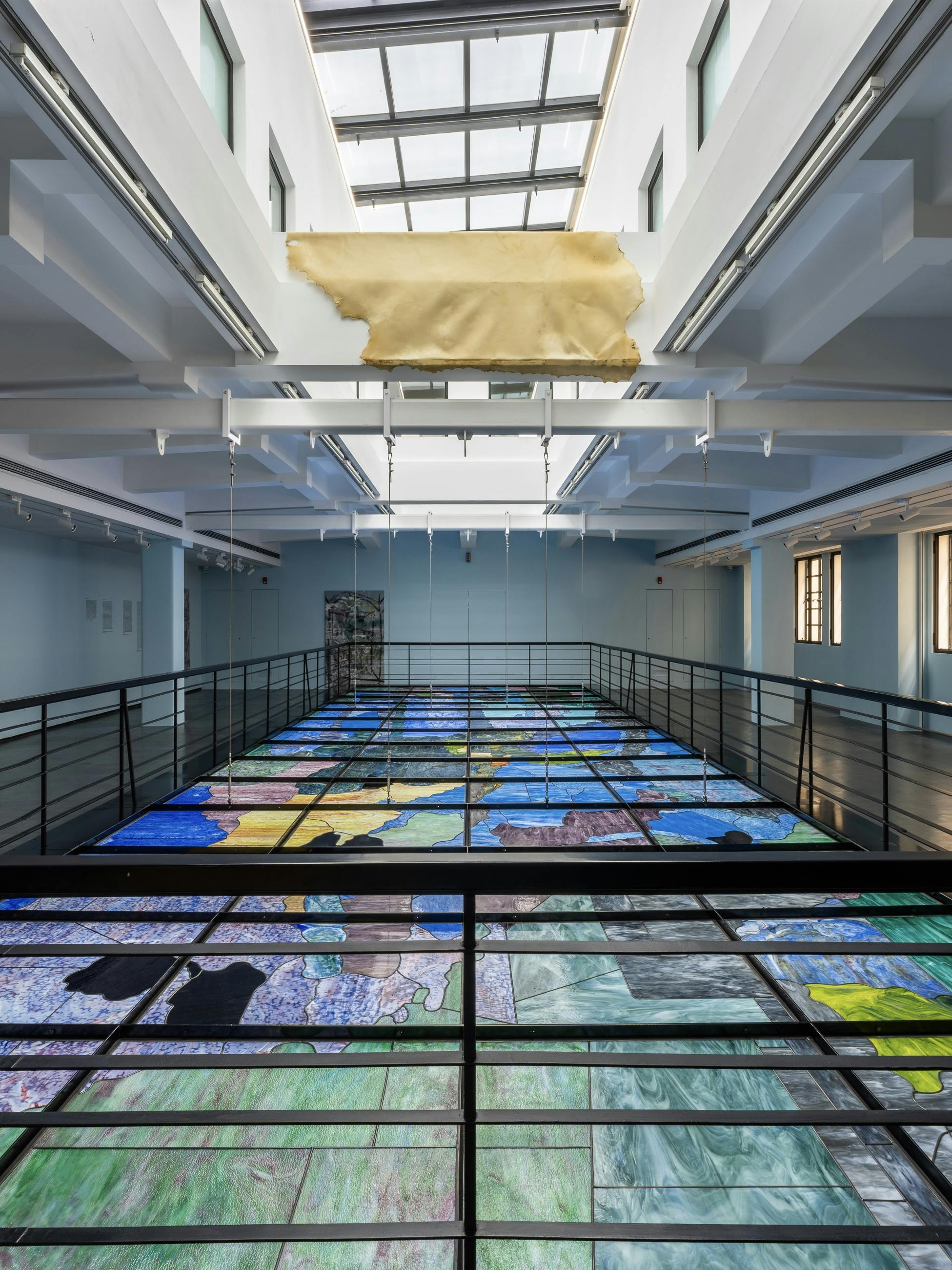 A rawhide hangs on a pilar between two floors, a railing divides the bottom part of the image, behind the railing is a stained glass ceiling, further back in the space is a painting, the space is painted baby blue.