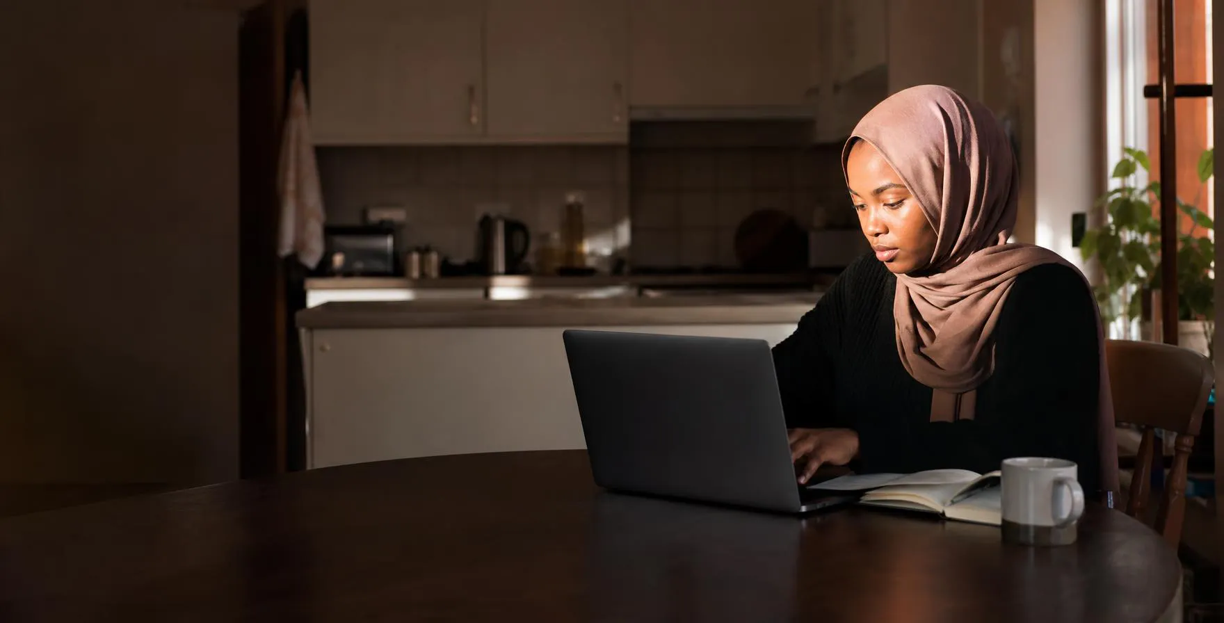 A young woman wearing a hijab working at a laptop at a kitchen table in morning light