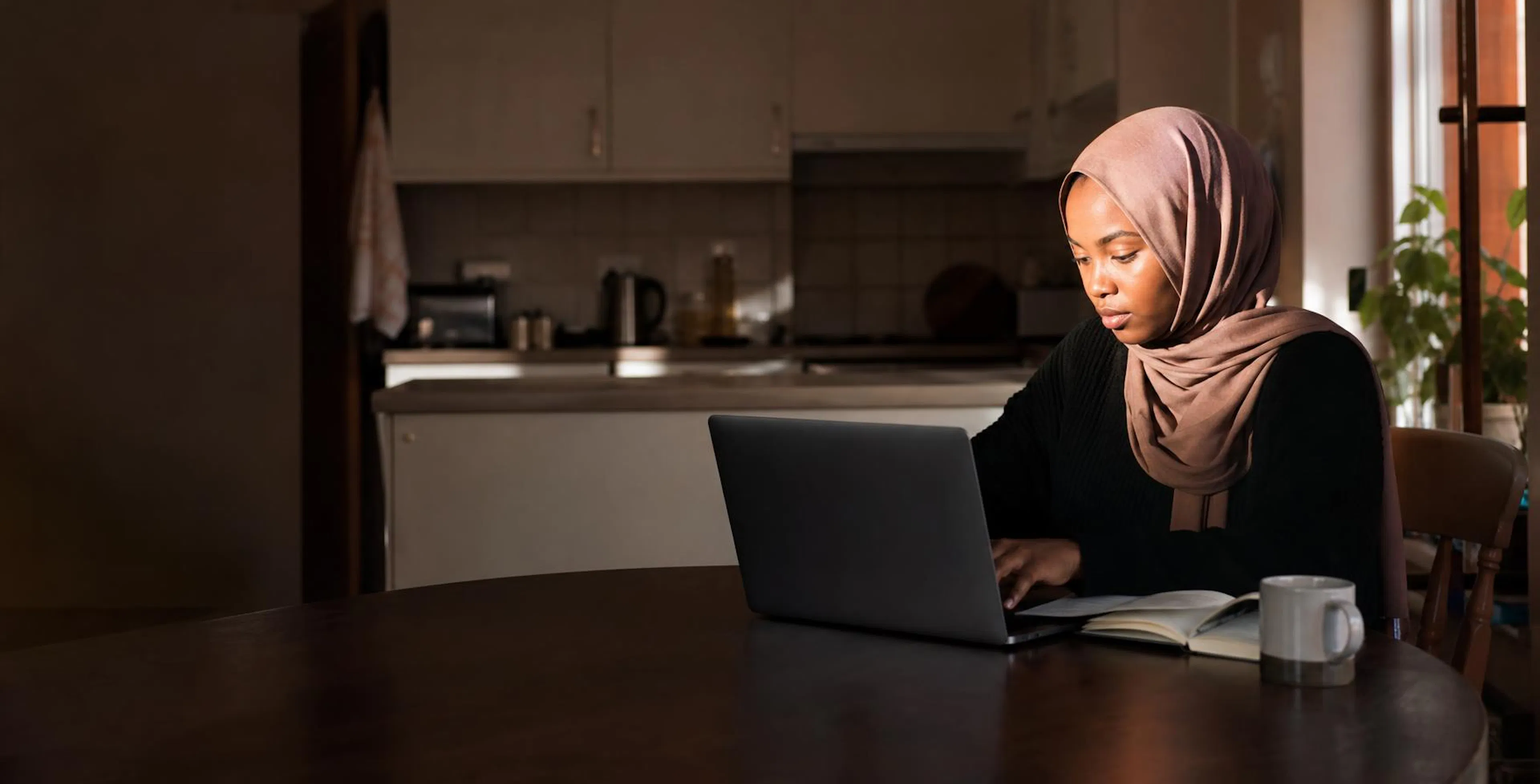 A young woman wearing a hijab working at a laptop at a kitchen table in morning light