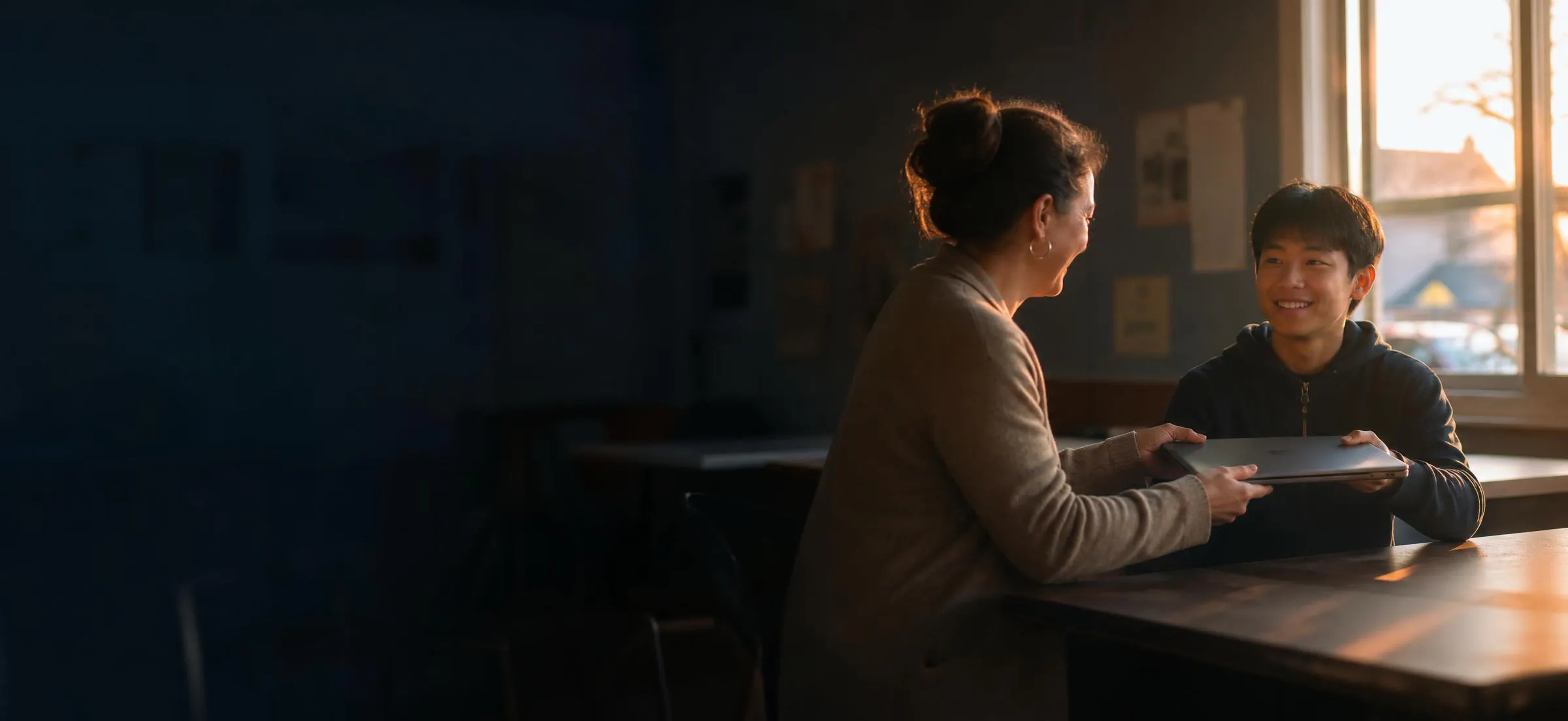 A woman handing a laptop to a smiling young man by a sunrise window