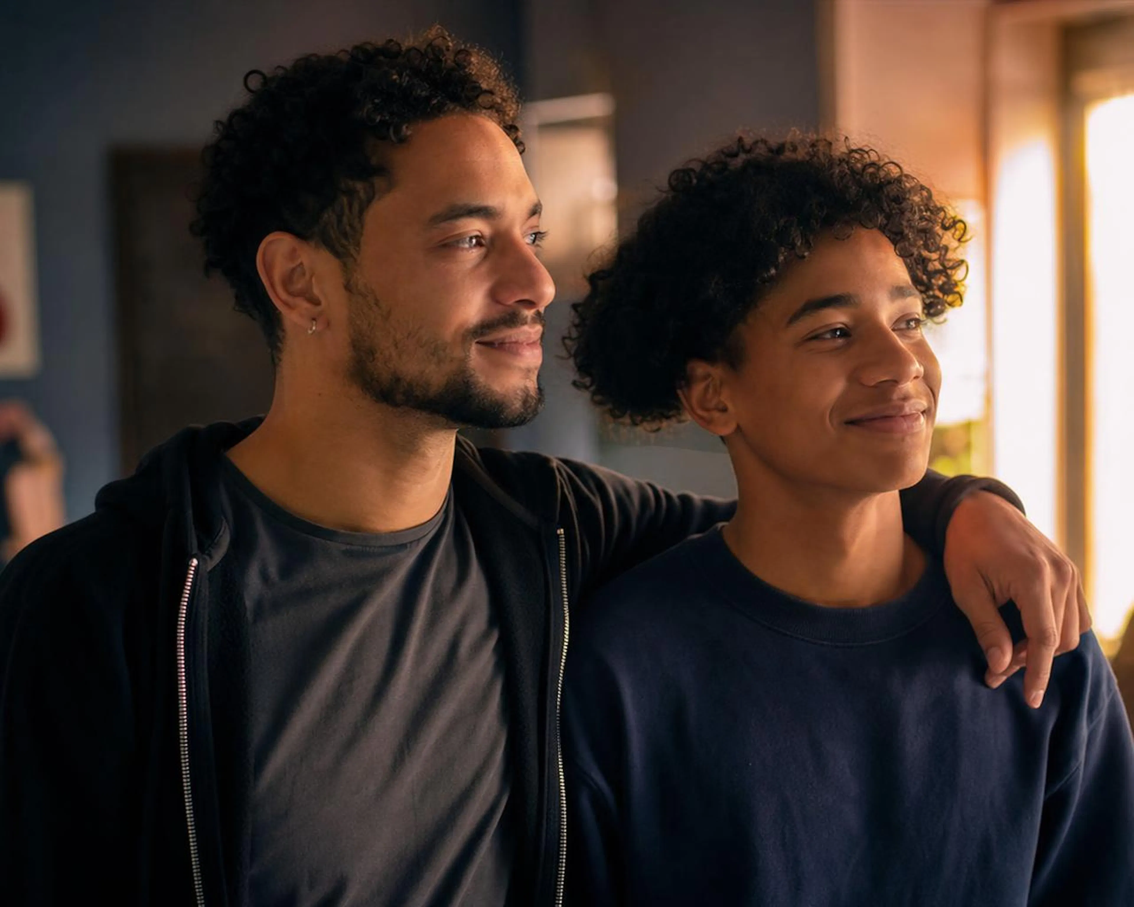 An older brother with his arm around his younger brother, both smiling toward a window
