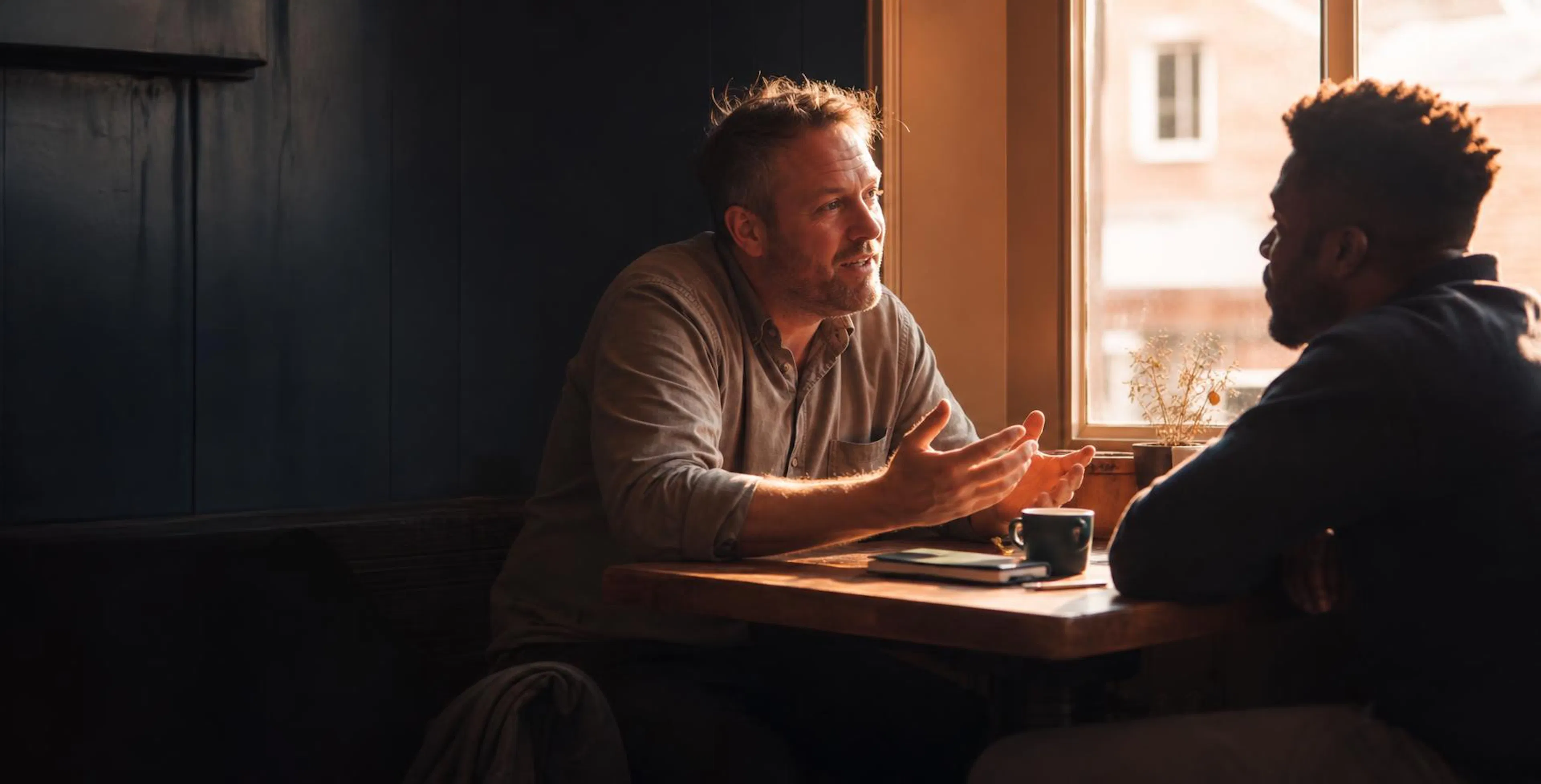 Two men talking across a small cafe table by a window