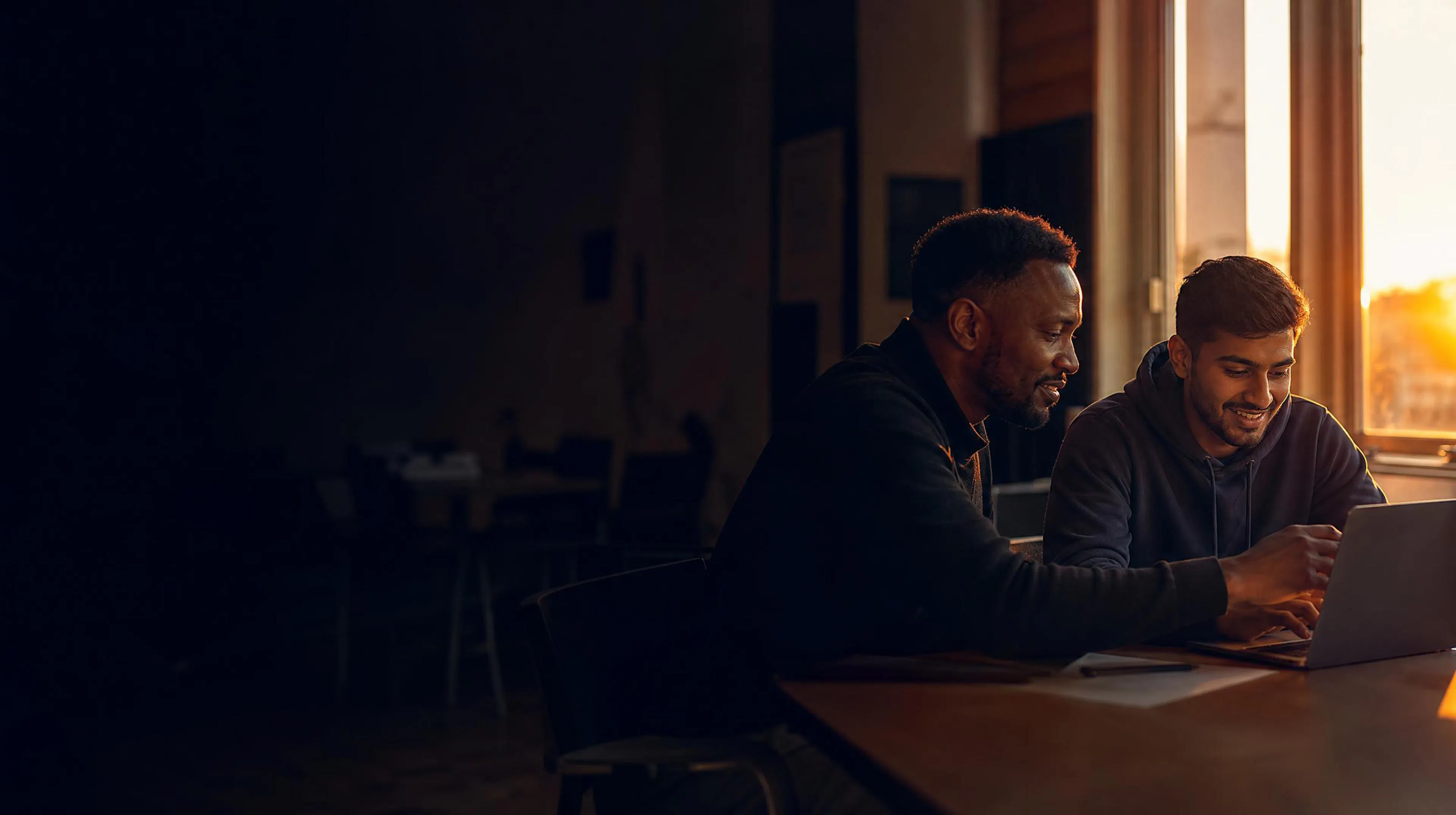 A mentor and a younger mentee smiling at a laptop by a window at sunrise