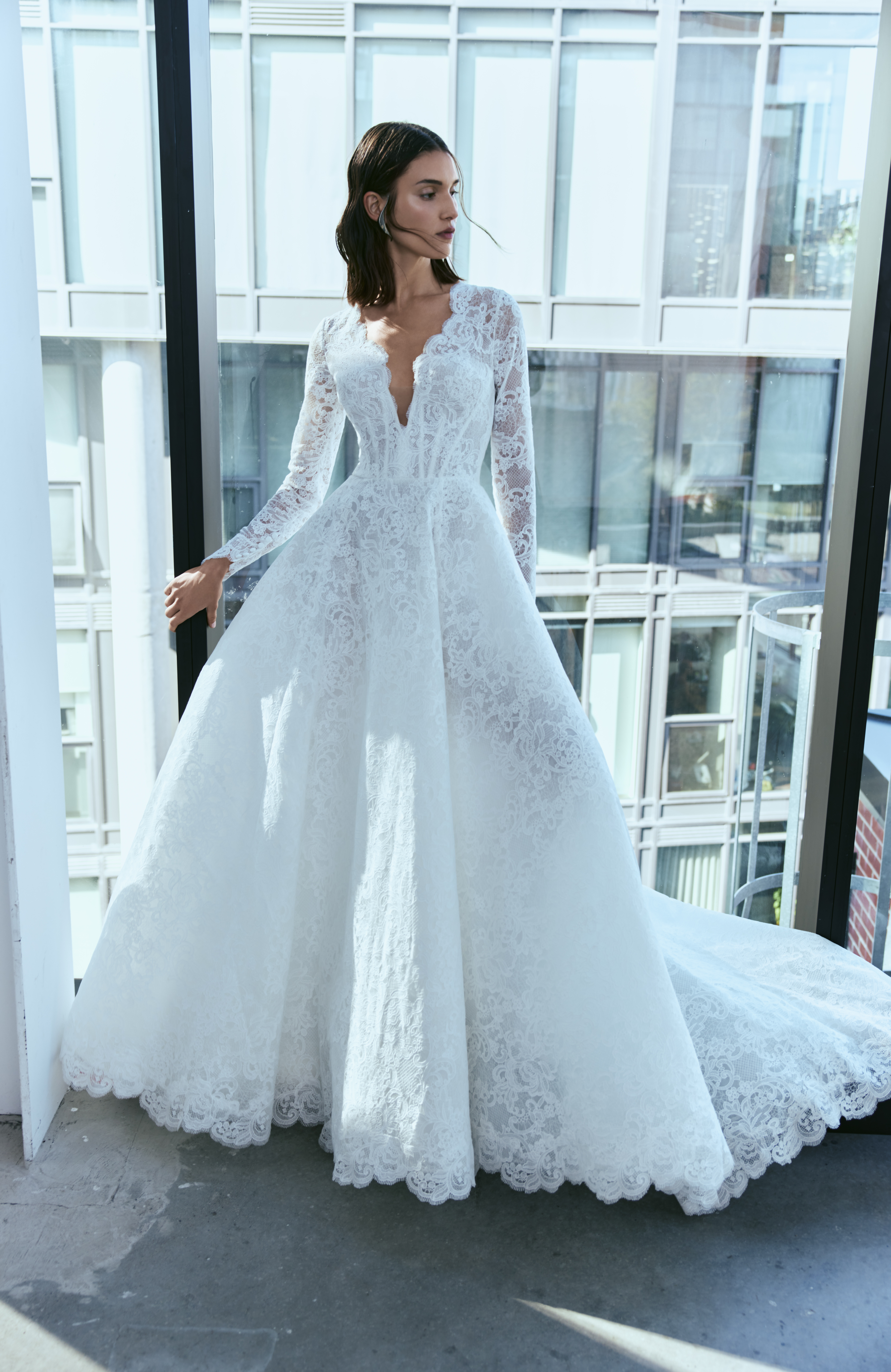Bride wearing the Aspen long-sleeve lace ballgown, standing indoors by large windows.