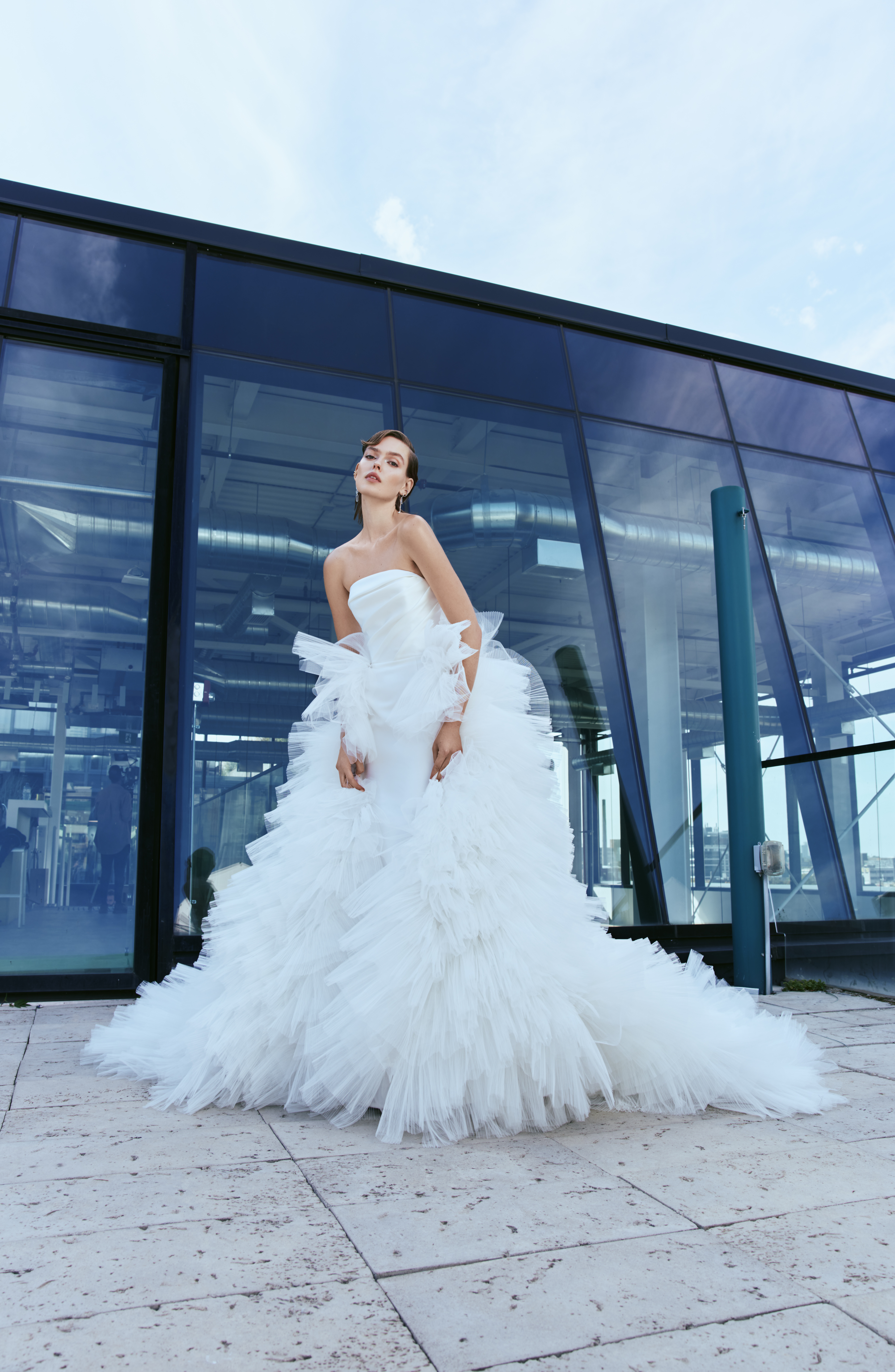 Parker RIVINI SS27 strapless fit-and-flare magnolia gown with a frothy hand-pleated tulle overskirt, shown outdoors against a modern architectural backdrop.