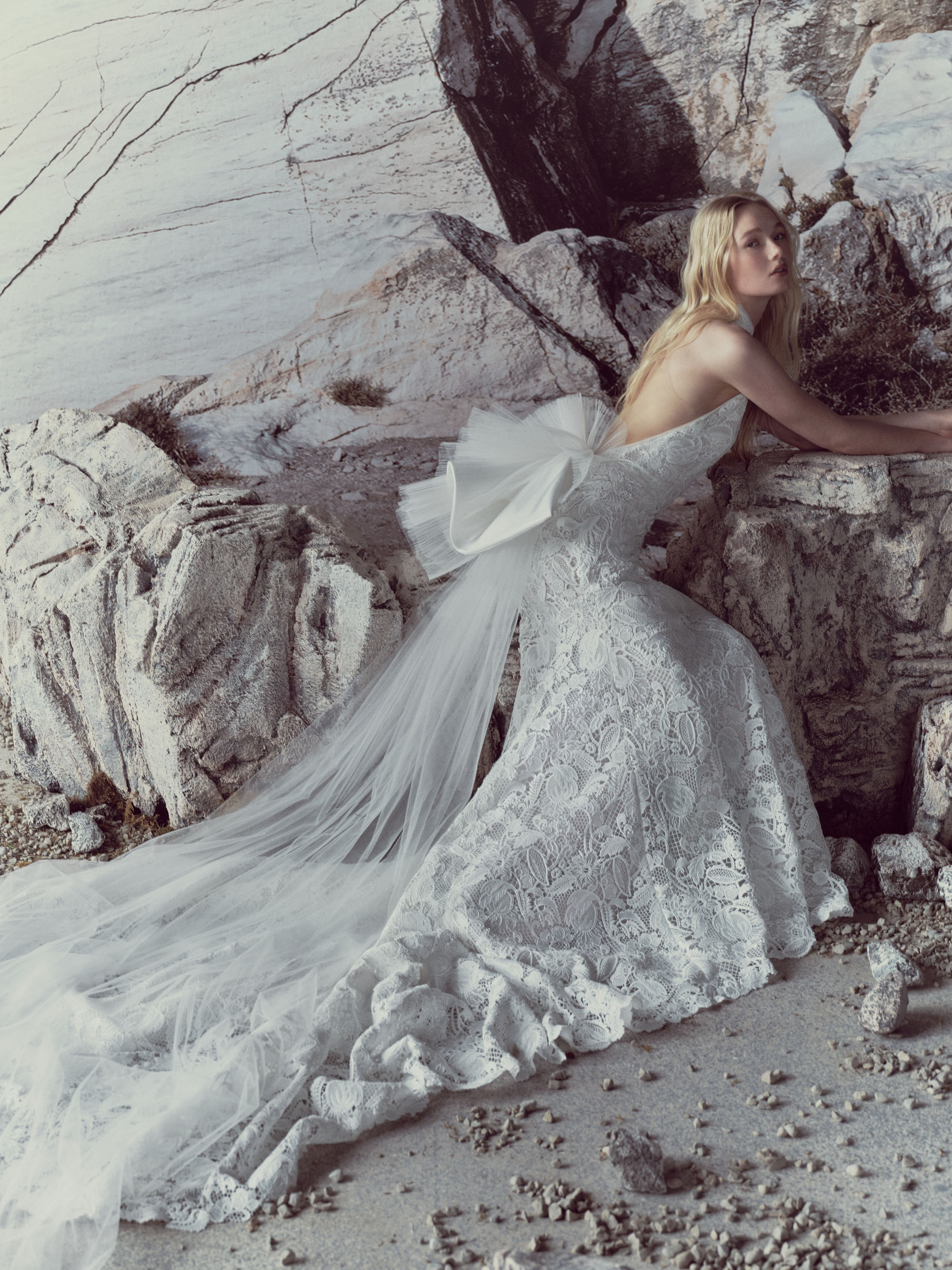 Sylvie RIVINI SS27 halter sheath gown in guipure lace with cascading layered ruffles and a voluminous waterfall train, shown seated.
