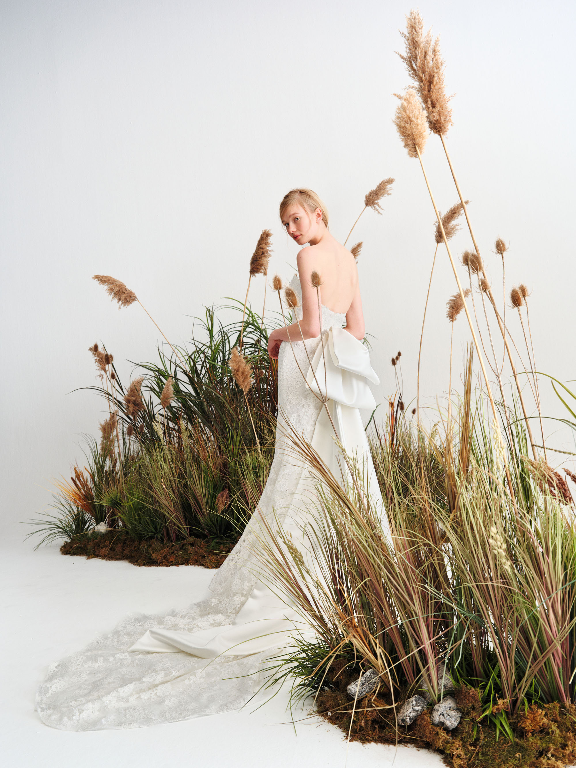 Full view of Cara showing the fitted lace silhouette, softly flared skirt, and flowing train among tall grasses.