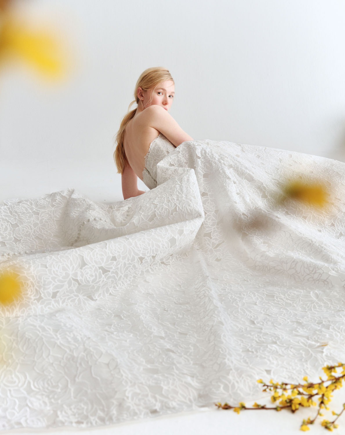 Sloane Alyne SS27 strapless ball gown in laser-cut embroidered organza with micro-pleated tulle at the neckline, shown seated.