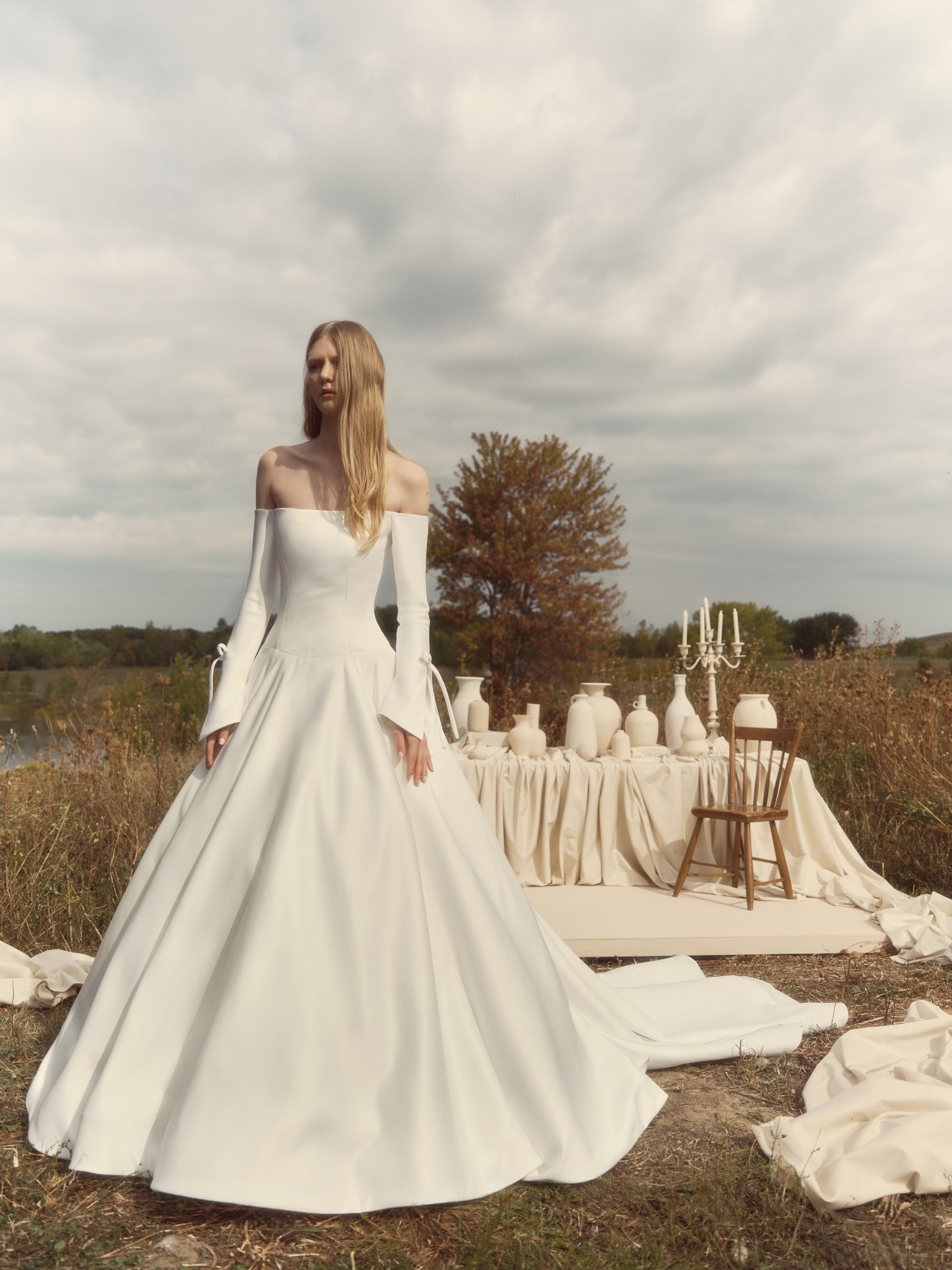 Grace off-the-shoulder ball gown with voluminous skirt, long tailored sleeves, pockets, and ribbon bow details, shown from the front.