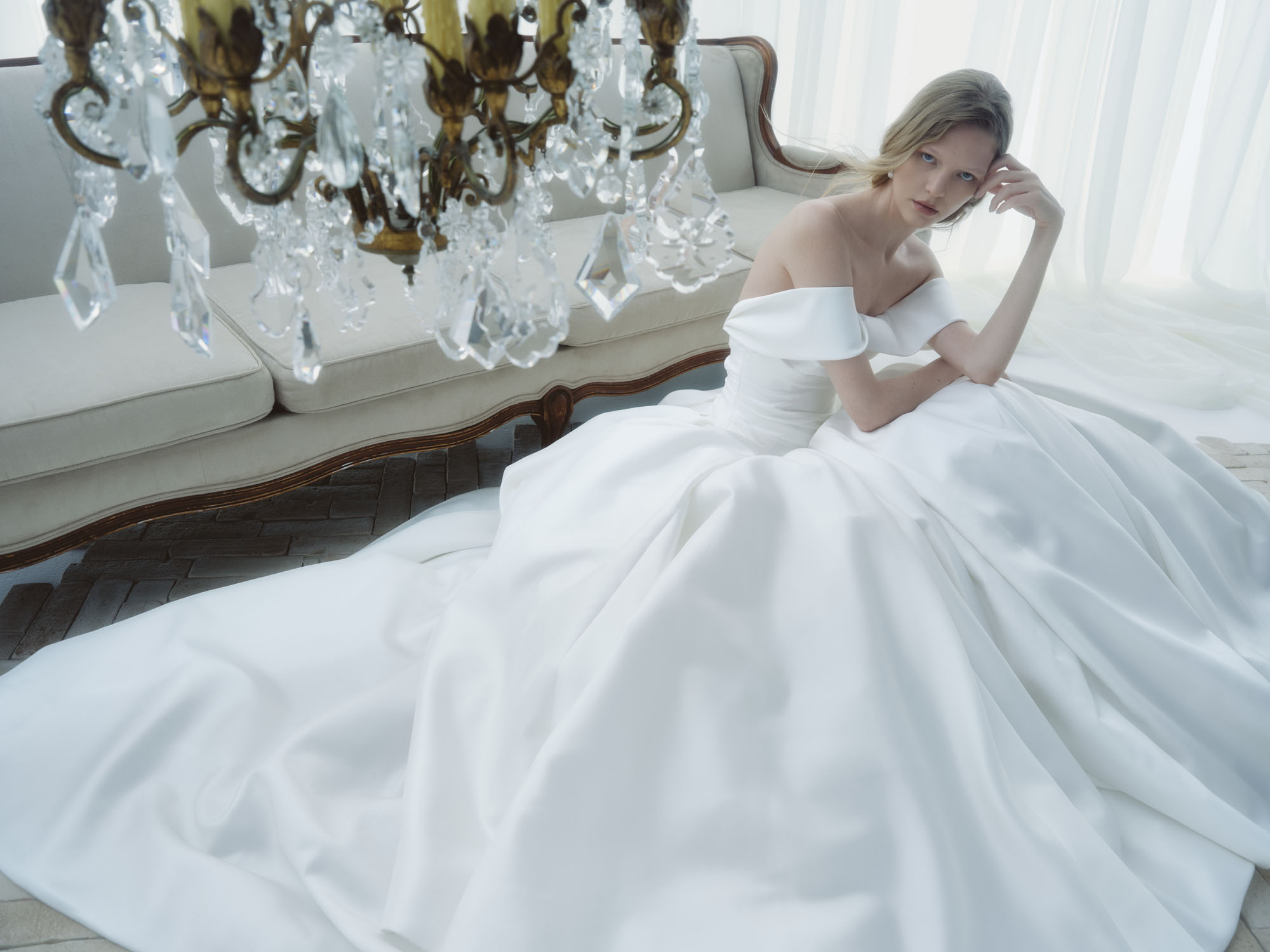 Aura RIVINI FW26 lustrous ball gown with draped bodice and twisted off-the-shoulder portrait collar, shown seated with the skirt spread around her.