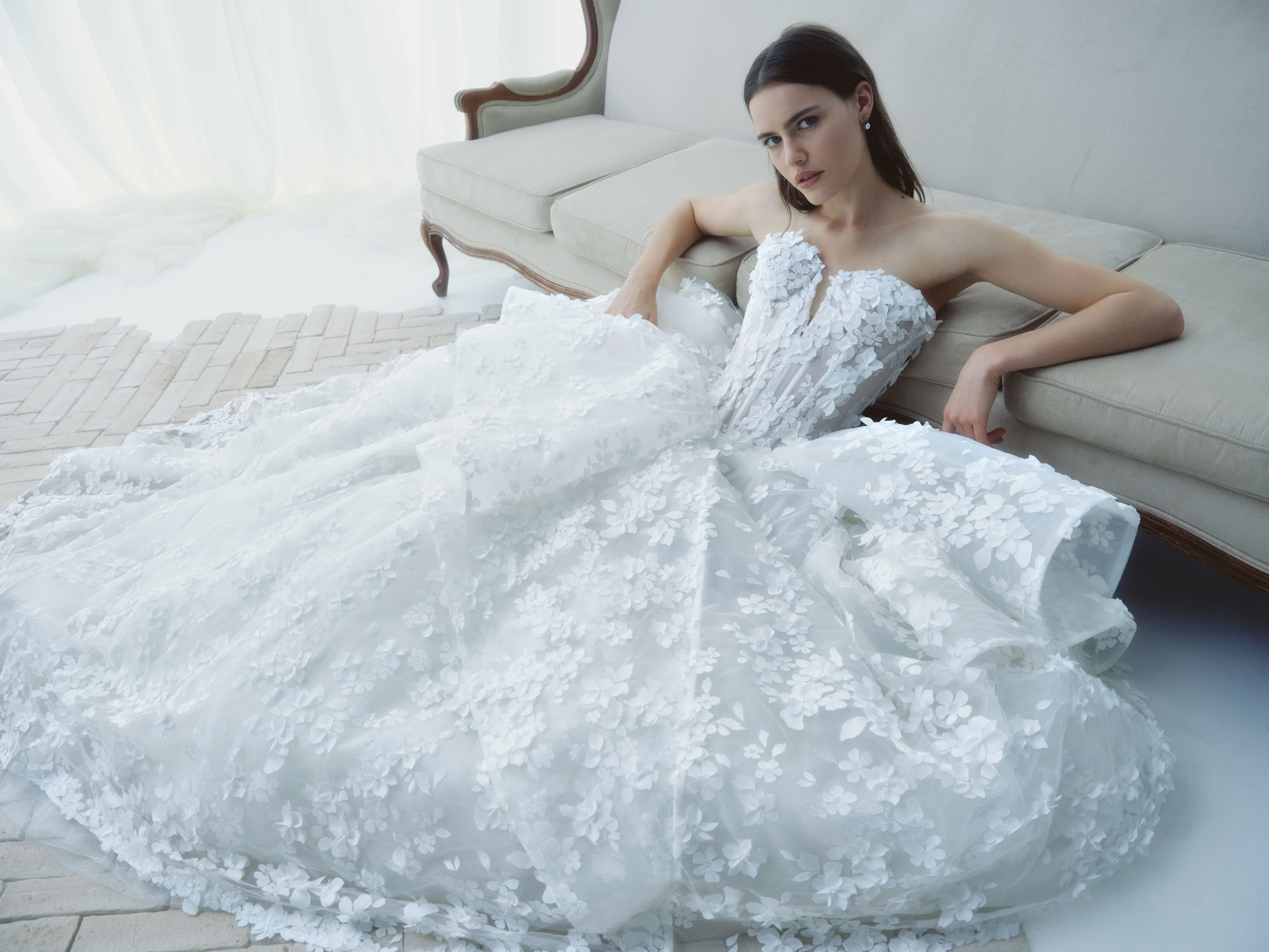 Reverie RIVINI FW26 strapless ball gown in shimmering 3D organza floral lace with a corseted bodice and layered peplum, shown seated.