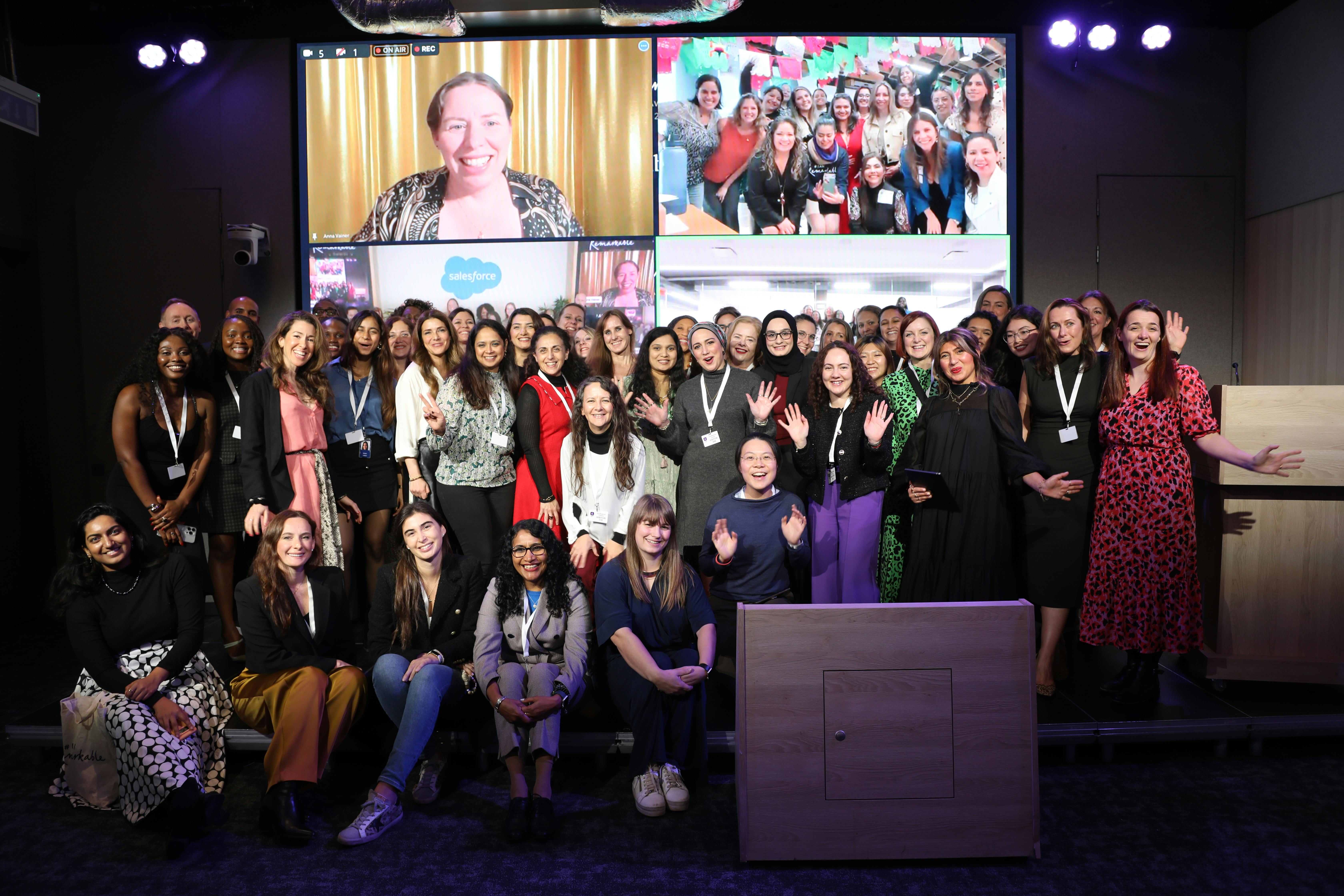 A diverse group of around 50 awards attendees stand on the stage in front of a screen which displays more participants dialling into the ceremony remotely. The mood is celebratory