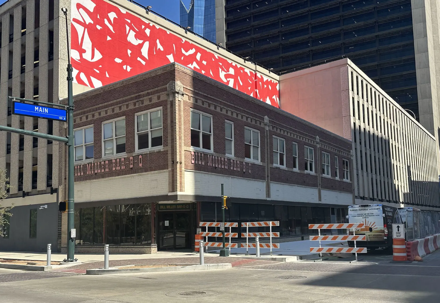 Two-storey brick corner building at Main Avenue with Bill Miller Bar-B-Q lettering across its facade, a red mural above it and downtown towers behind