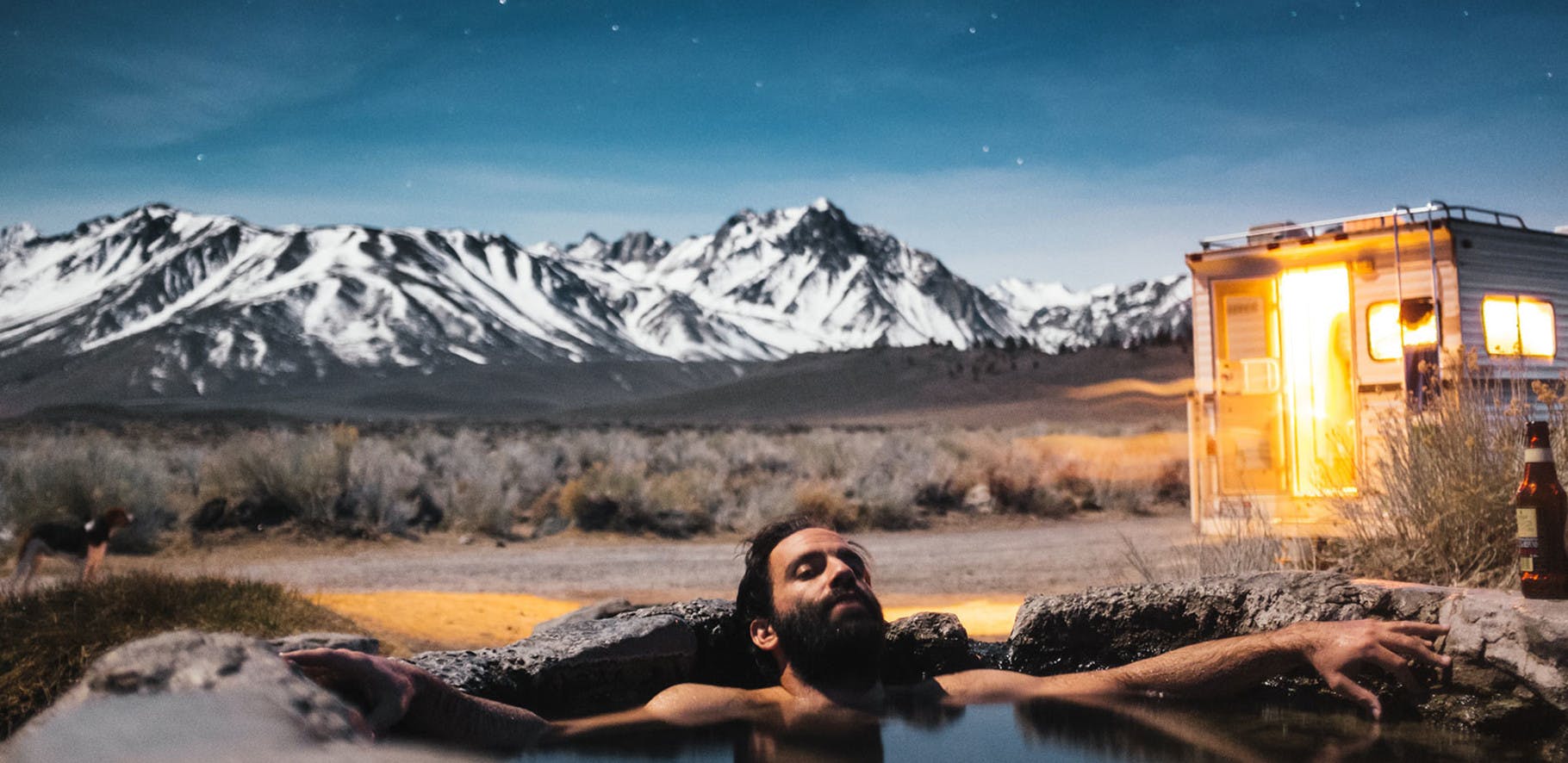 Man relaxing in an natural hot spring with his camper and a mountain range in the background