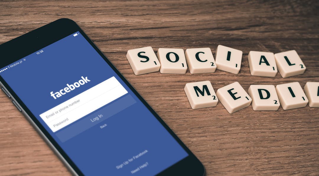 A phone with facebook on the screen is sitting on a desk with the words social media spelled out in scrabble titles sitting next to it