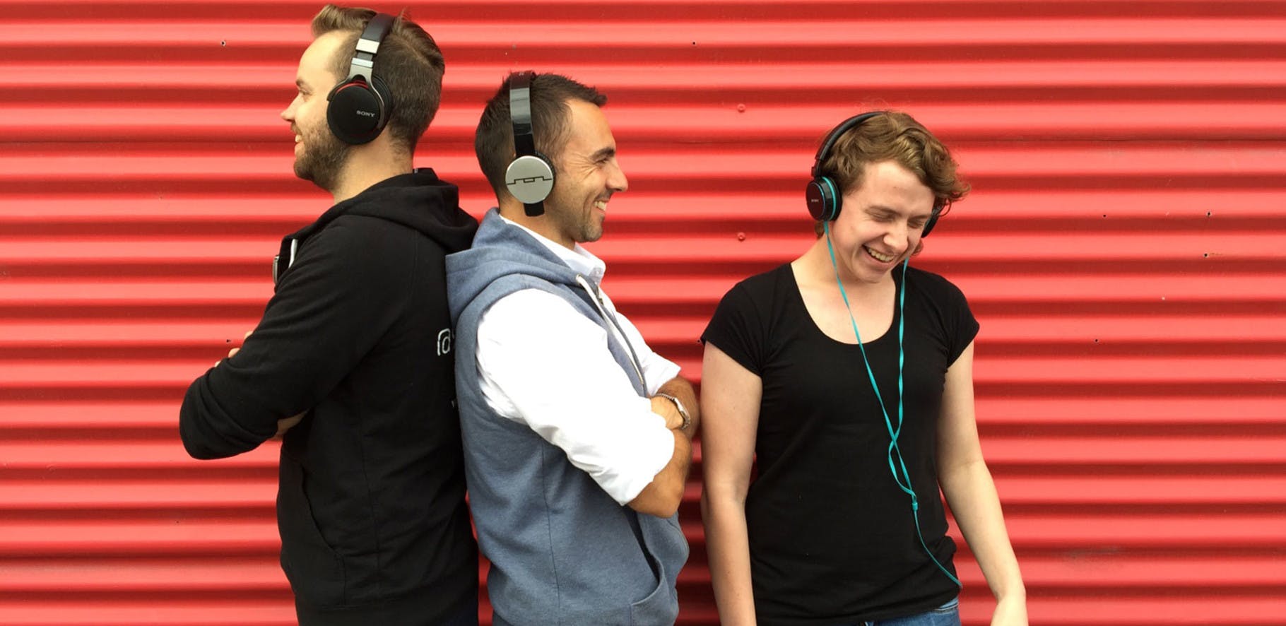 three people wearing headphones are smiling and laughing whilst standing against a bright red corrugated metal background