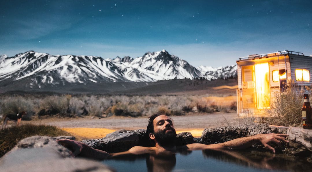 Man relaxing in an natural hot spring with his camper and a mountain range in the background