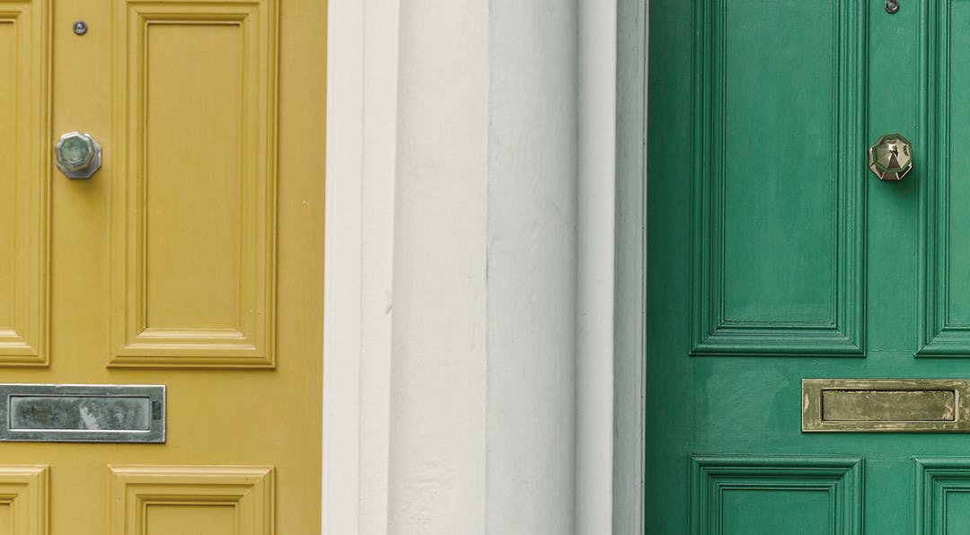 two doors are seperated by a white wall, there is a yellow door on the left and a green door on the right.