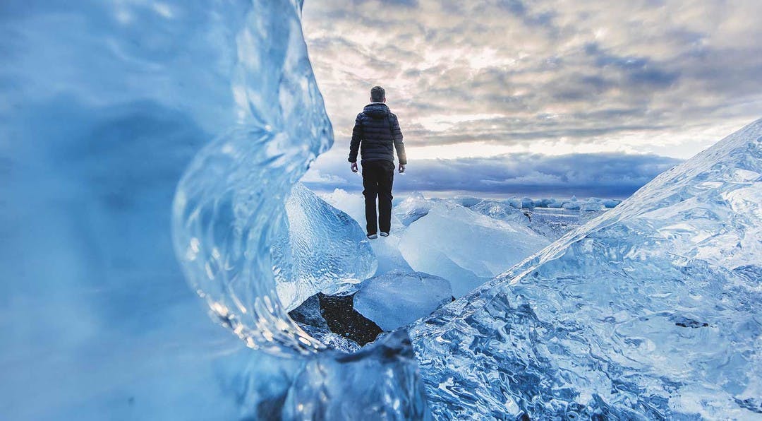 close up image looking through glacier ice, with a man standing in the background looking out over the vast glacier