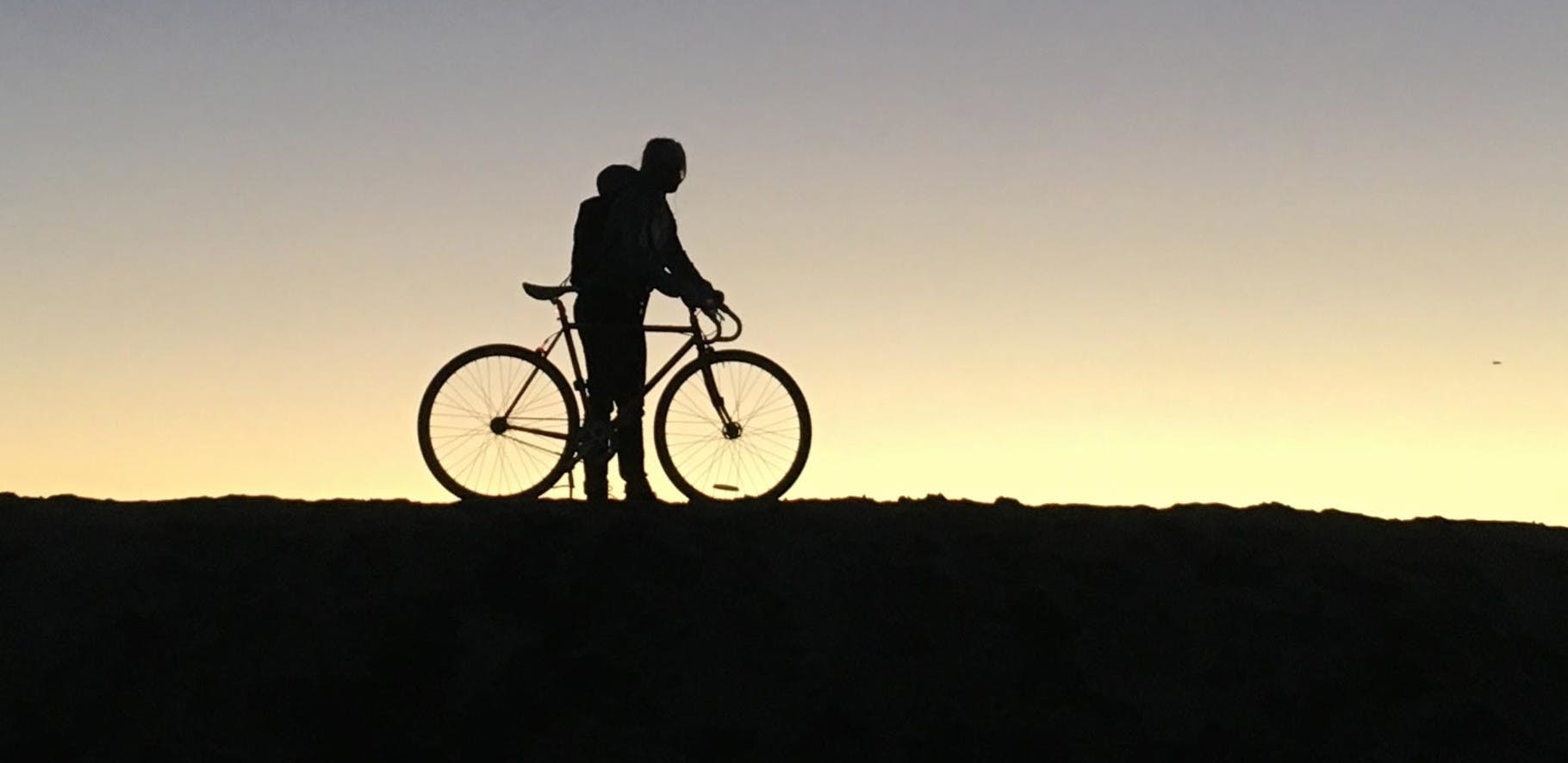 Black silhouette of a man and his bicycle against an evening sunset backdrop
