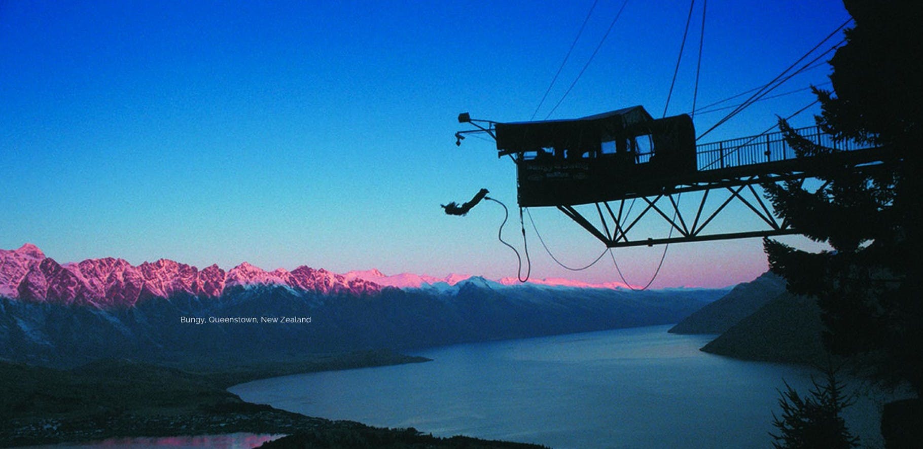 Black silhouette of a person bungy jumping from a tower, over looking a beautiful mountain range and lake at dusk.