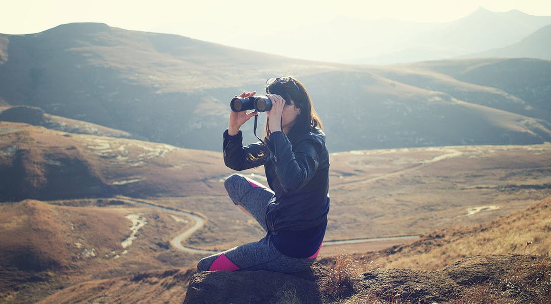 woman using binoculars sitting on a hill with the valley behind her in the background