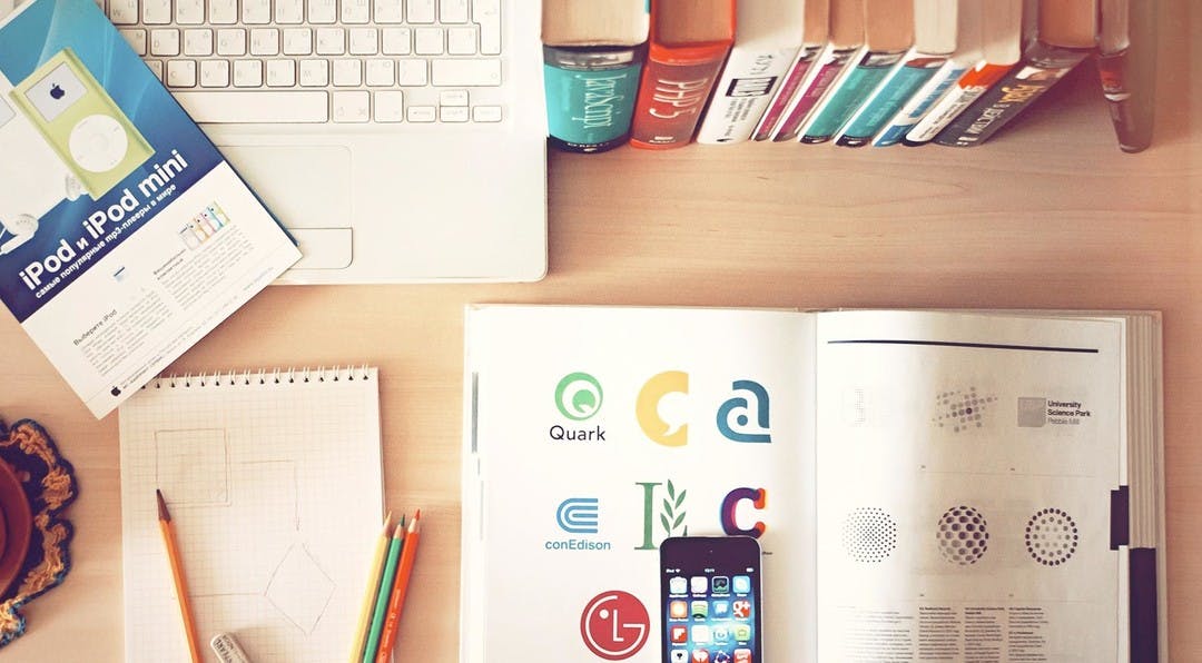 looking down onto an office desk which shows a lap, notepad, pencils, phone and an open book