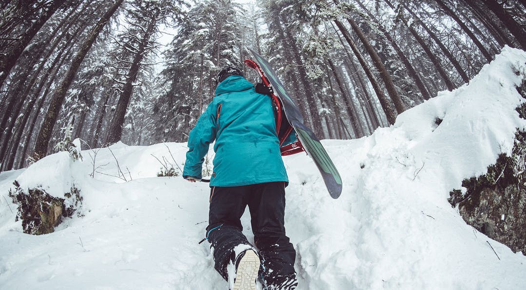 snowboarder carrying his board on his back tramping through the snow amongst the trees