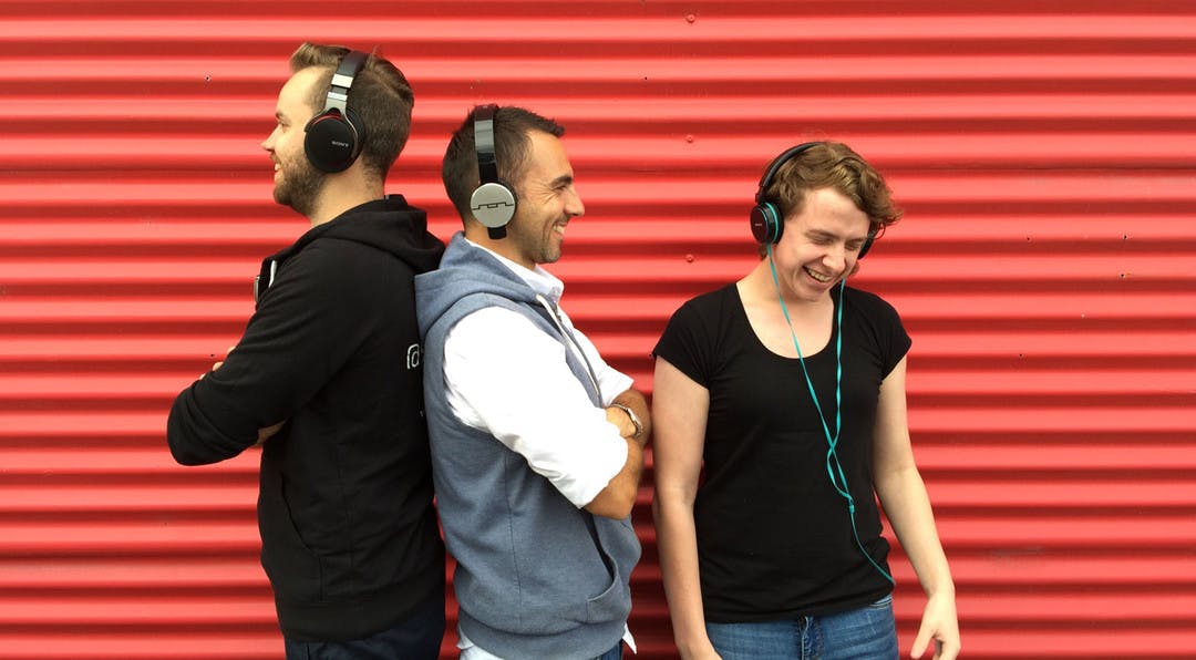 three people wearing headphones are smiling and laughing whilst standing against a bright red corrugated metal background