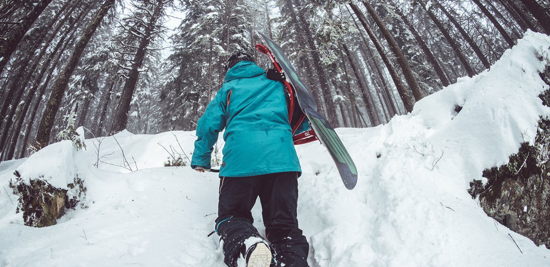 snowboarder carrying his board on his back tramping through the snow amongst the trees