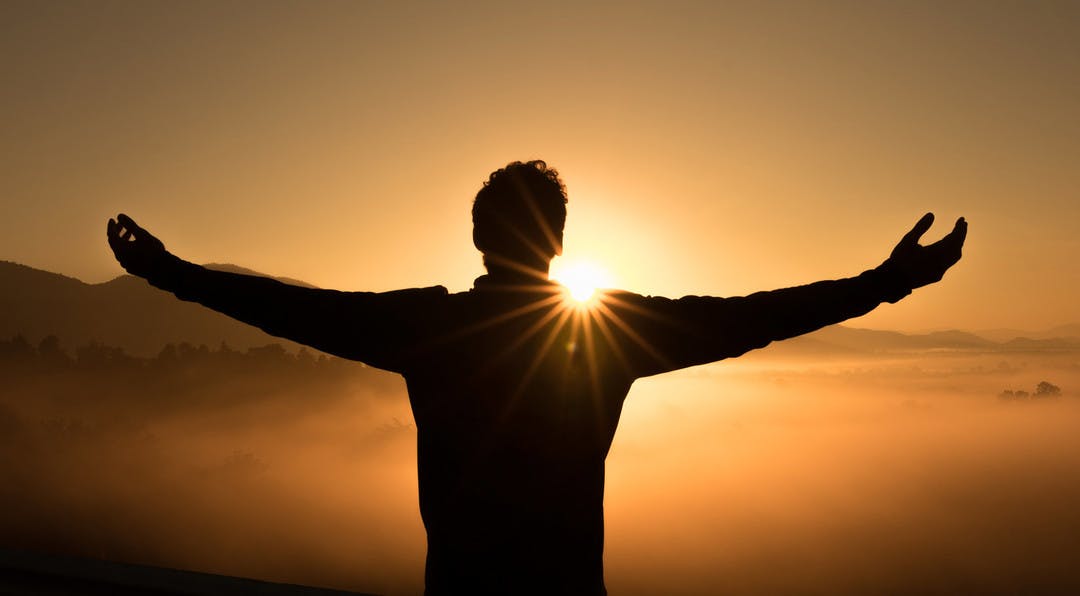 Black silhouette of a man standing on a high peak with arms outstretched overlooking mountains and mist at sunset.