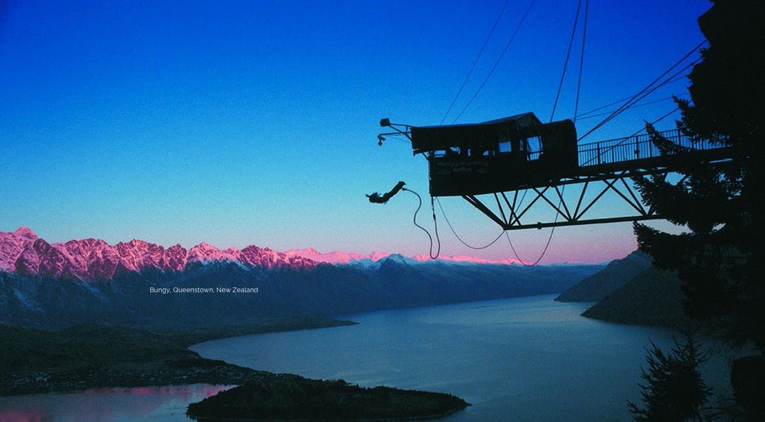 Black silhouette of a person bungy jumping from a tower, over looking a beautiful mountain range and lake at dusk.