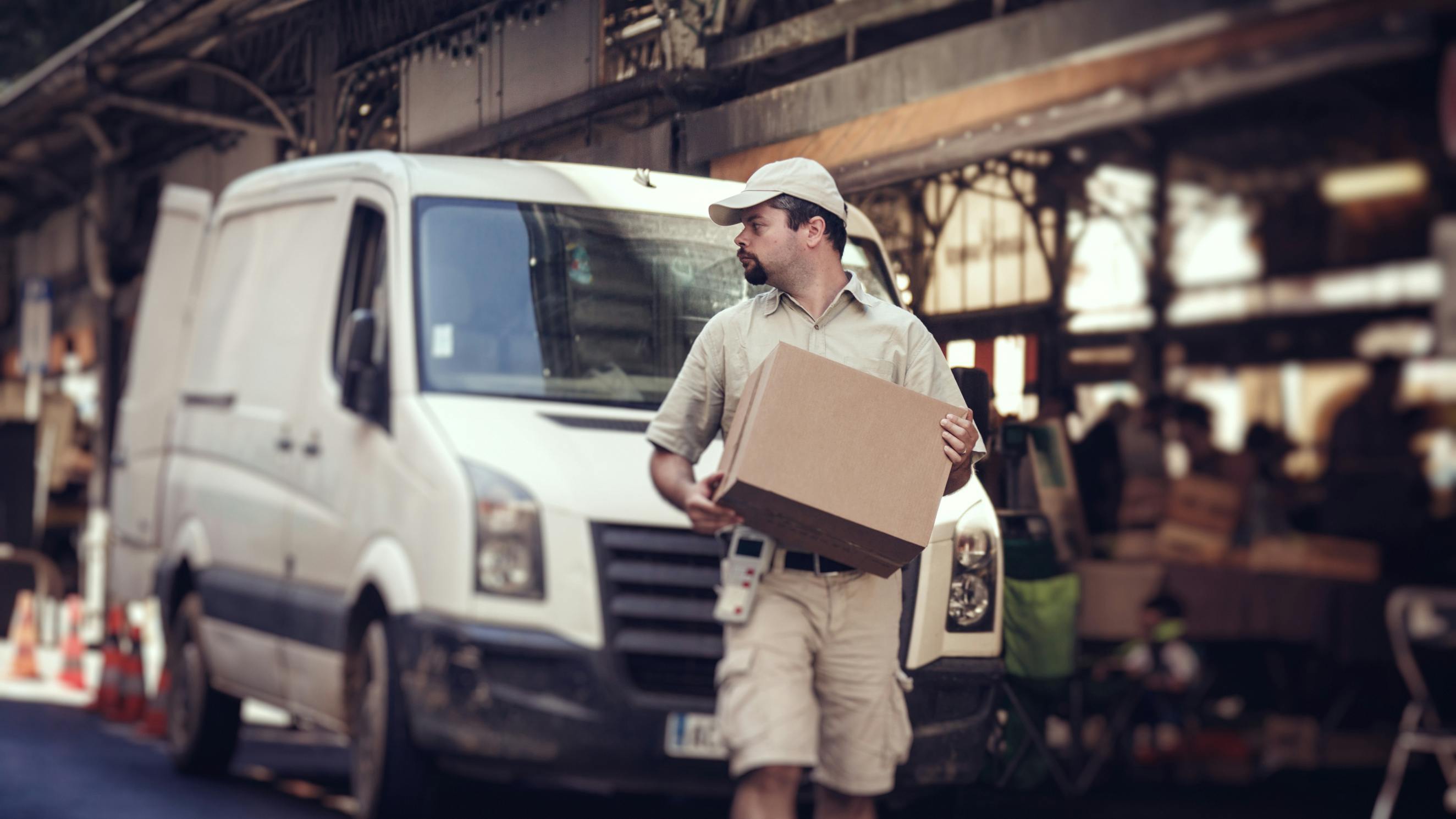 Delivery person with package in front of white van on a busy street with shops and pedestrians in the background.