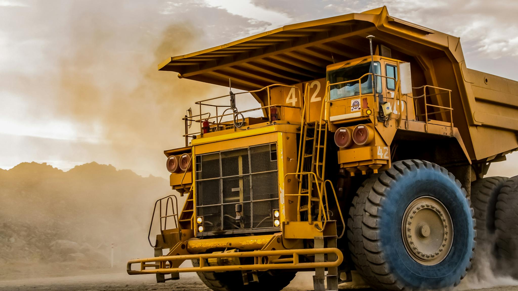 Yellow mining dump truck #42 driving on a dusty road with mountains in the background.