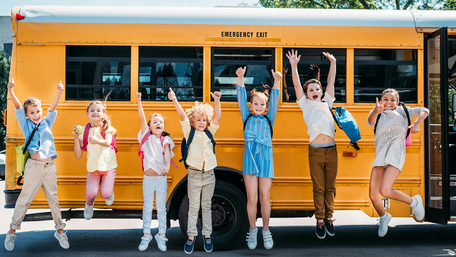 Children jump in front of a yellow NYC school bus with visible emergency exit sign, highlighting pedestrian safety.