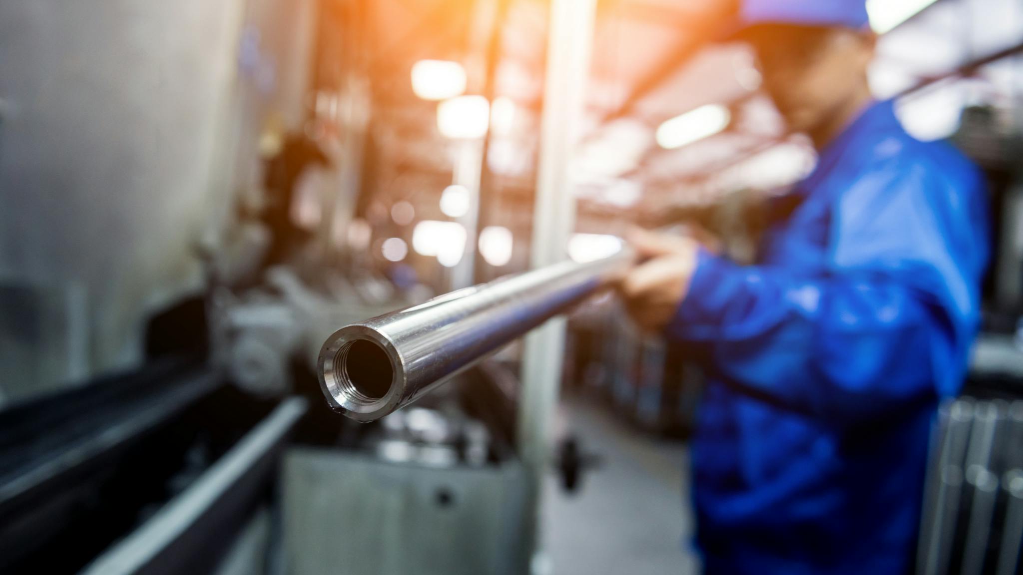Worker in blue uniform holds metal pipe in industrial area with blurred background.