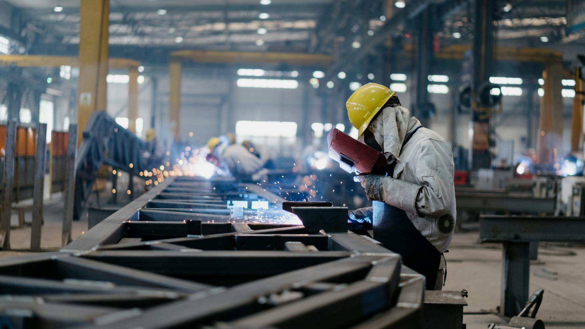 Workers welding in a factory with protective gear and machinery, showcasing industrial manufacturing operations.