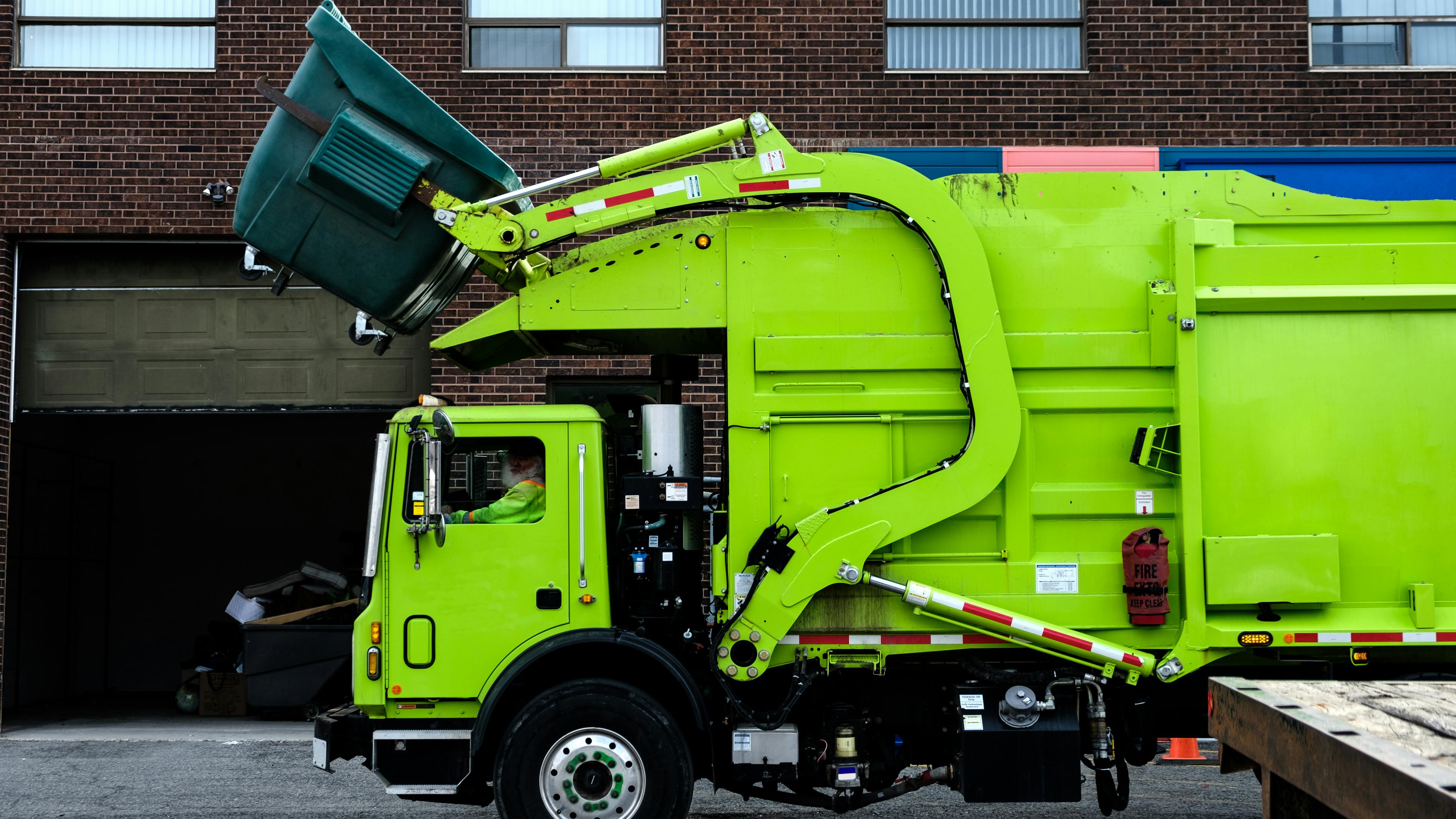 Green garbage truck lifting dumpster in front of brick building with garage door, showcasing waste collection.