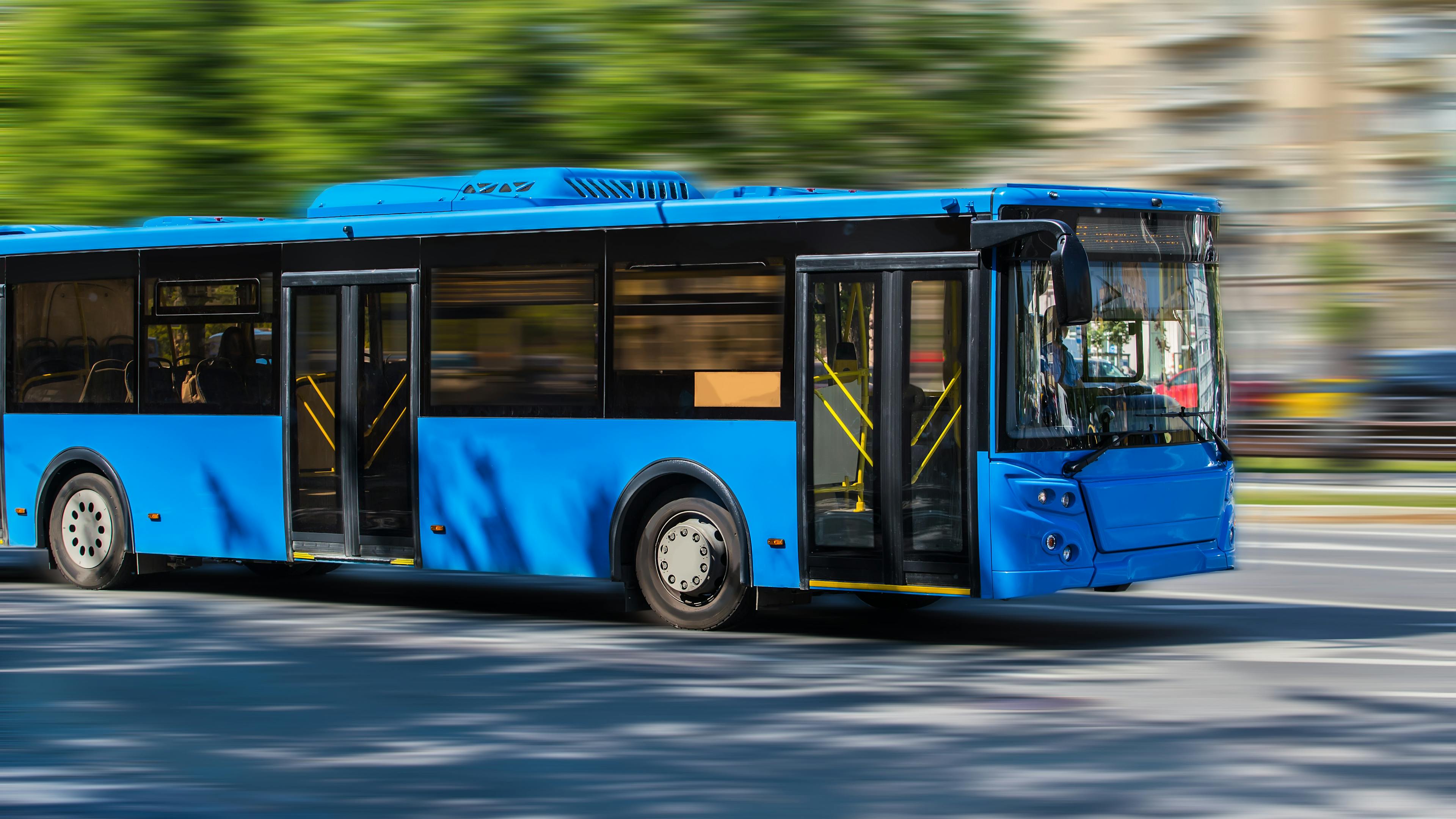 Blue city bus driving past blurred trees and buildings on a sunny street.