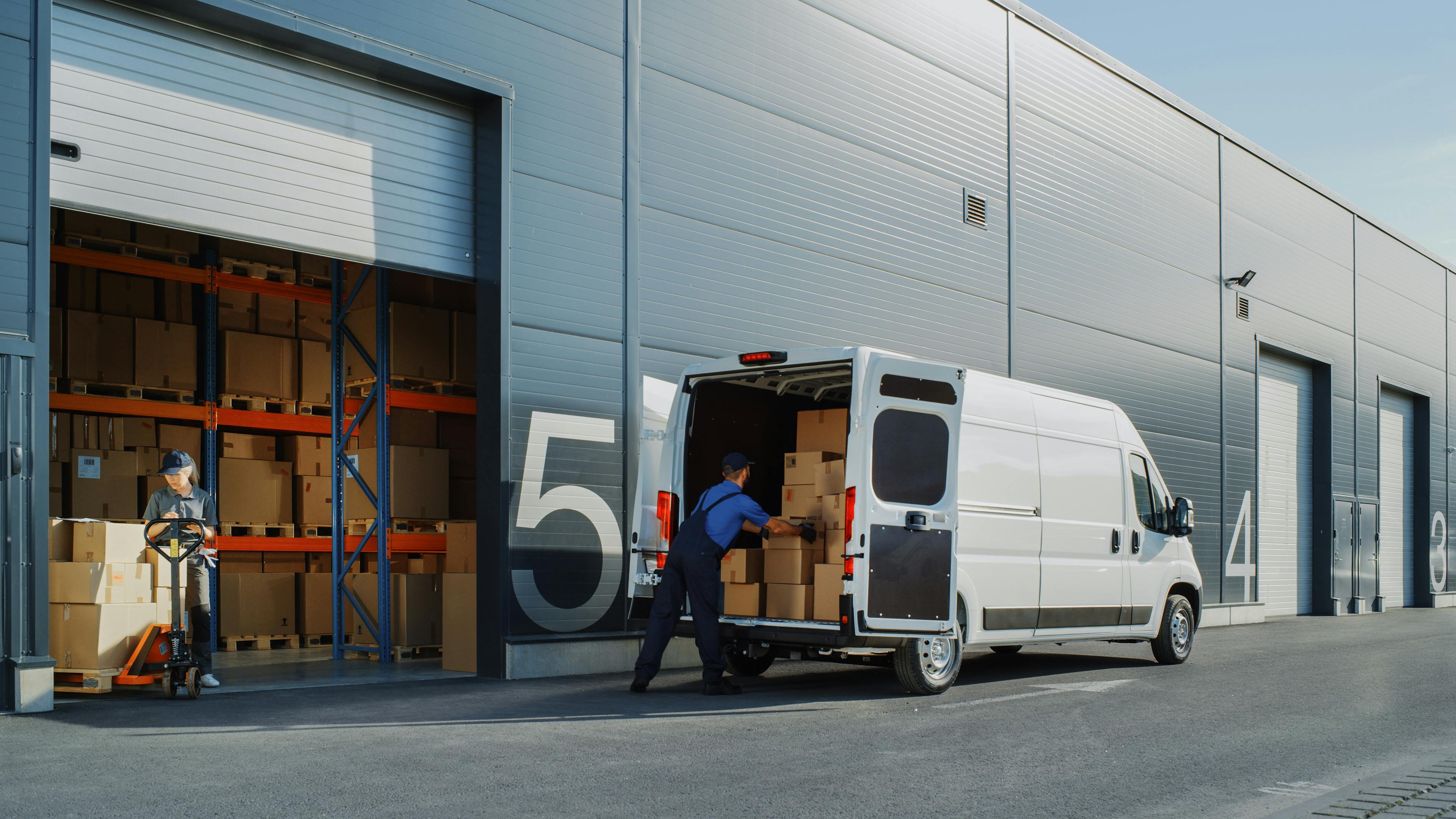 Warehouse workers load boxes into a white delivery van outside a large warehouse with numbered loading doors.