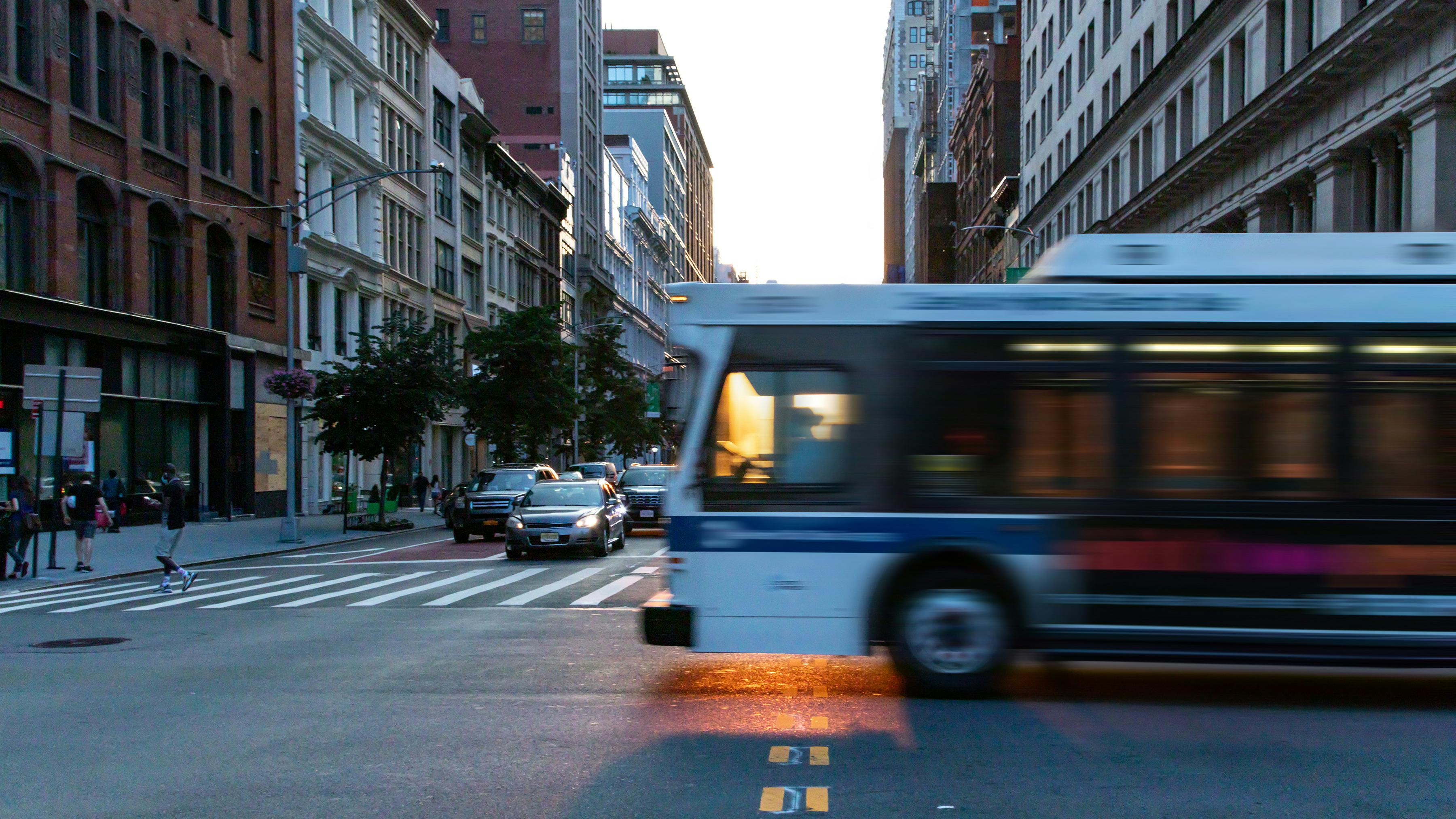 Bus, cars, and pedestrians move through a city intersection at sunset.