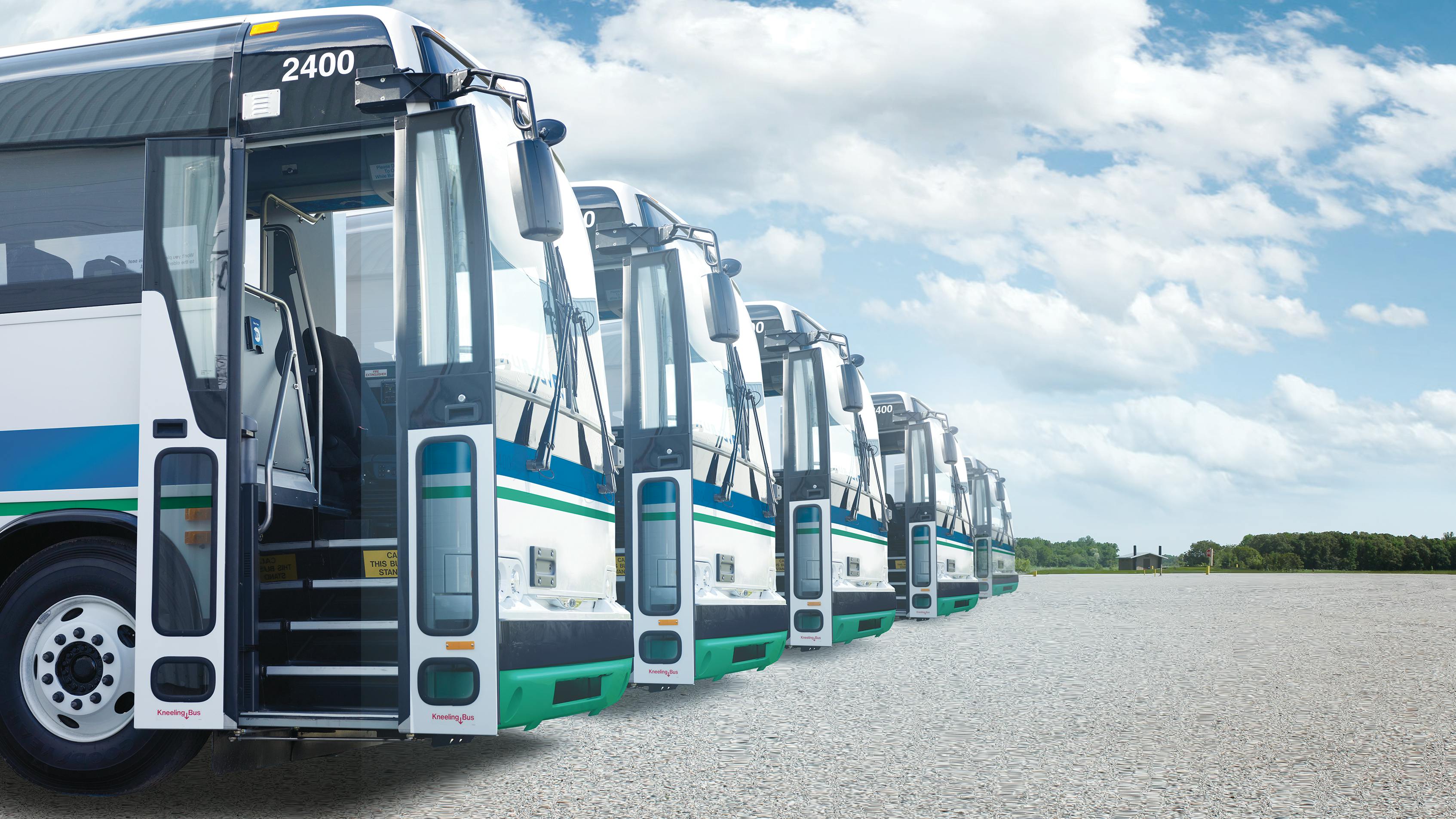 Five white and green buses parked on gravel lot beneath partly cloudy sky, highlighting fleet transportation.