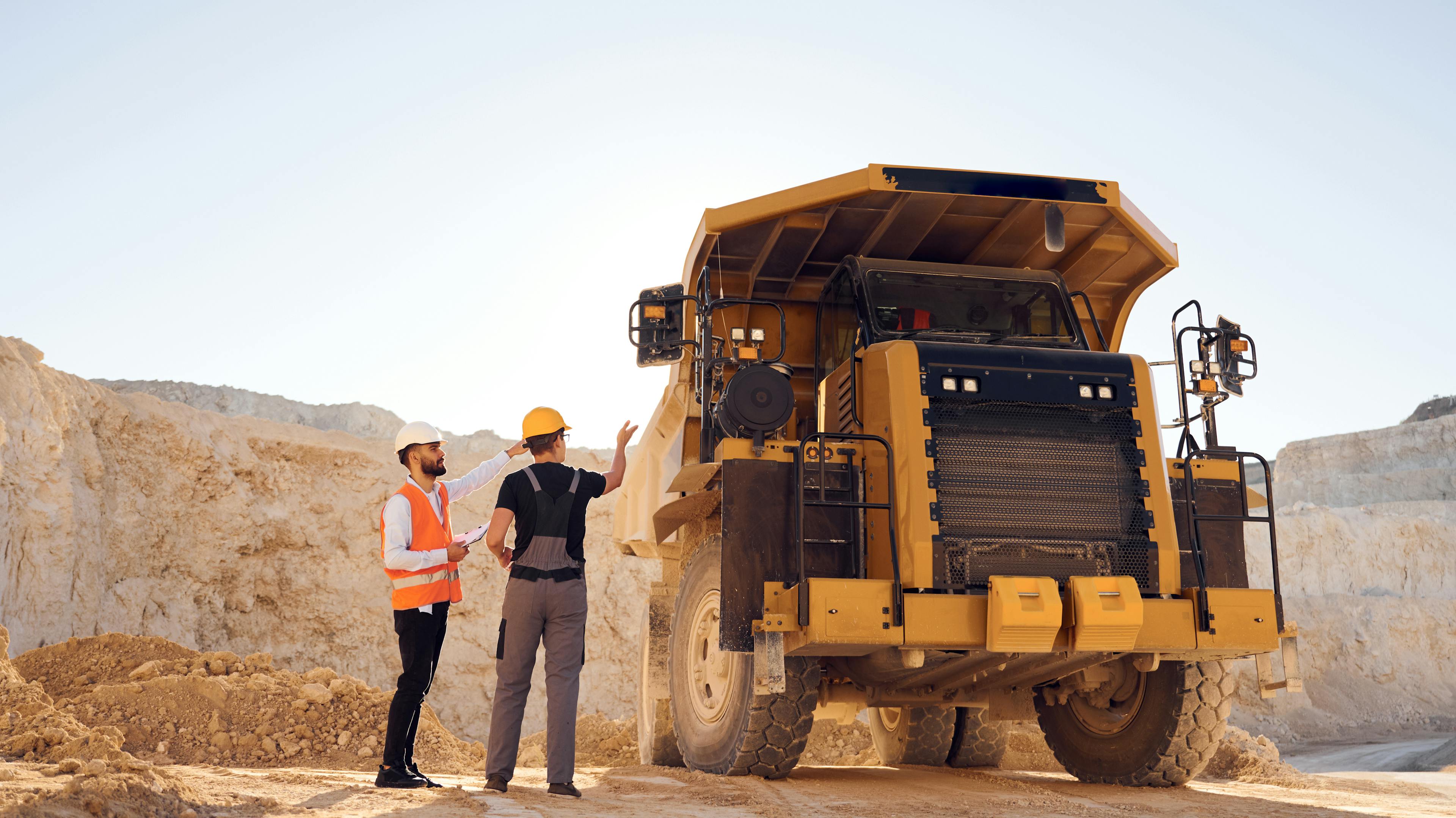 Two construction workers in safety gear beside a yellow dump truck at a quarry site.