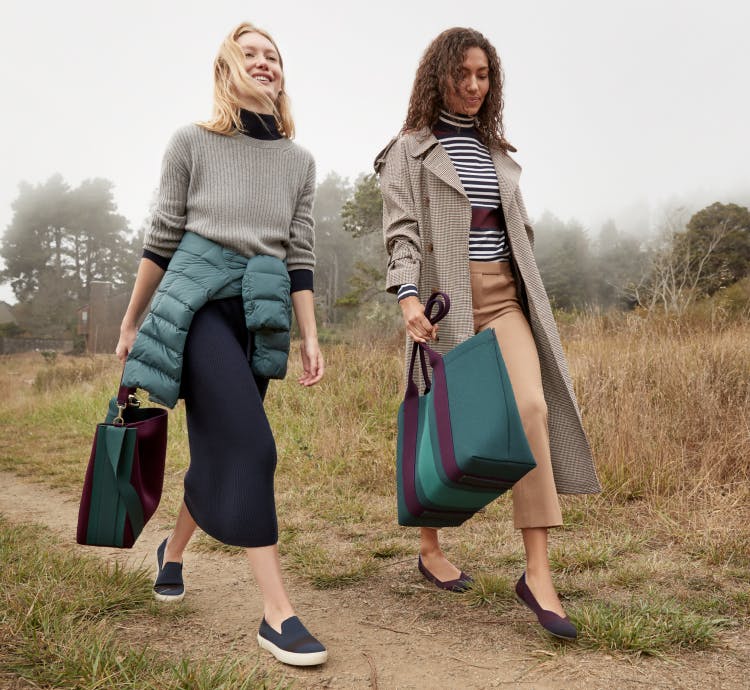 Two models holding The Bucket Bag in Dark Plum and The Essential Tote in Deep Spruce wearing The Sneaker in Night Stripe and The Flat in Blackberry Captoe.