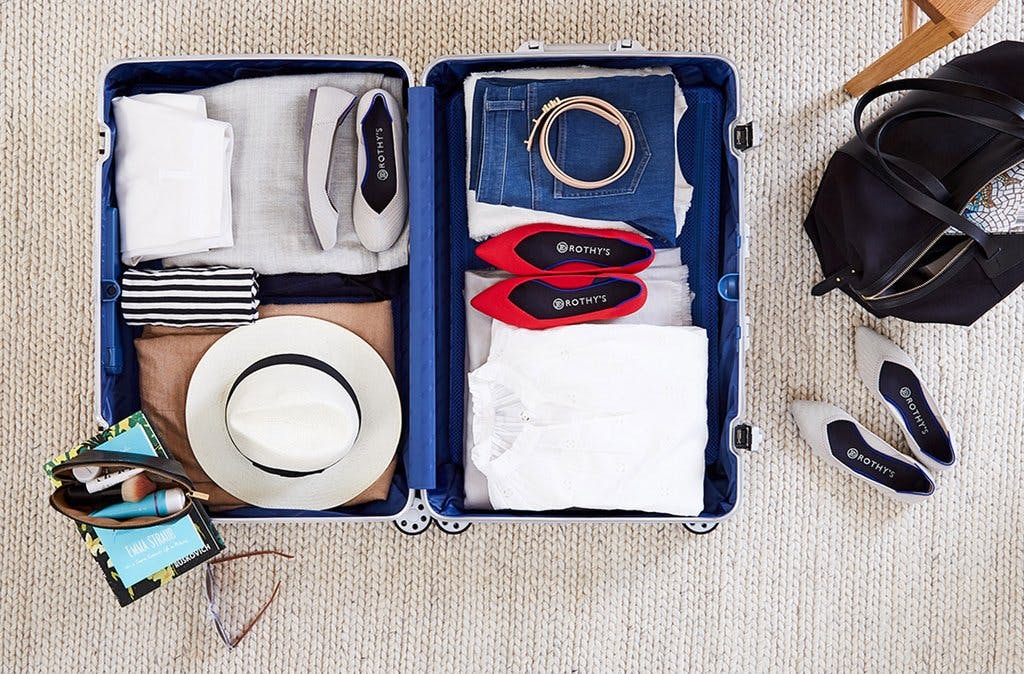 An open luggage with folded clothes, a hat, and two pairs of Rothy's flats. 