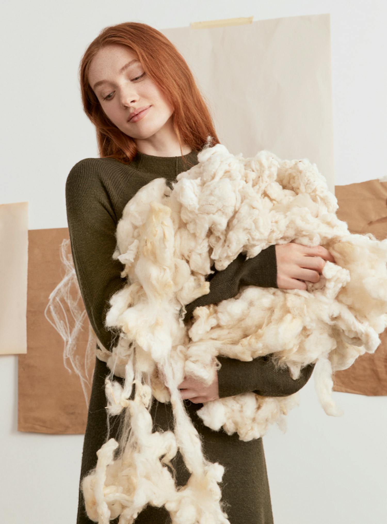 From left to right, model holding a bunch of wool, The Merino Flat in Bordeaux Herringbone shown on model’s feet and two plastic bottles shown standing on a white stool.